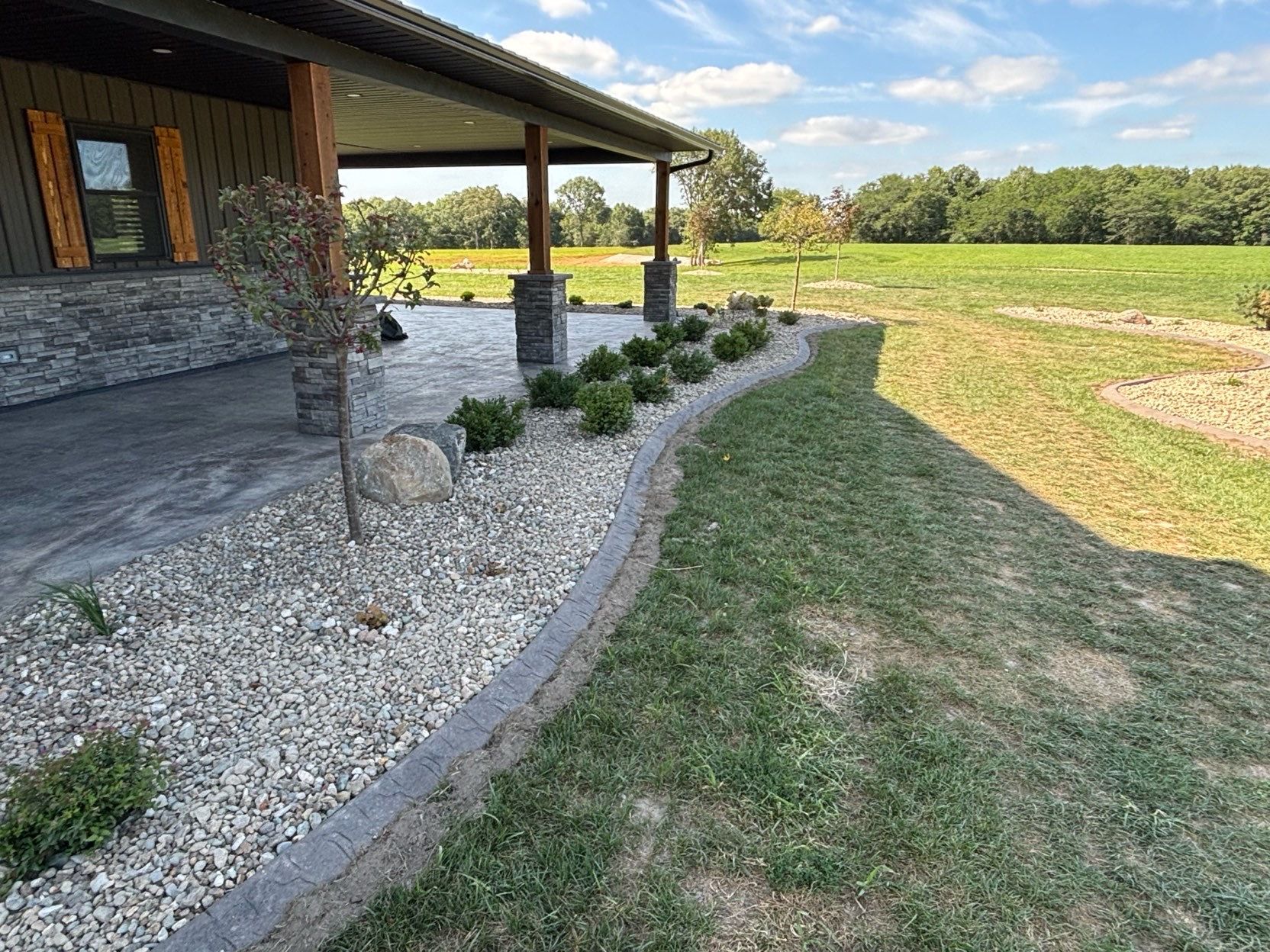 Stone-bordered gravel bed beside a covered porch, with small shrubs and a grassy yard beyond.