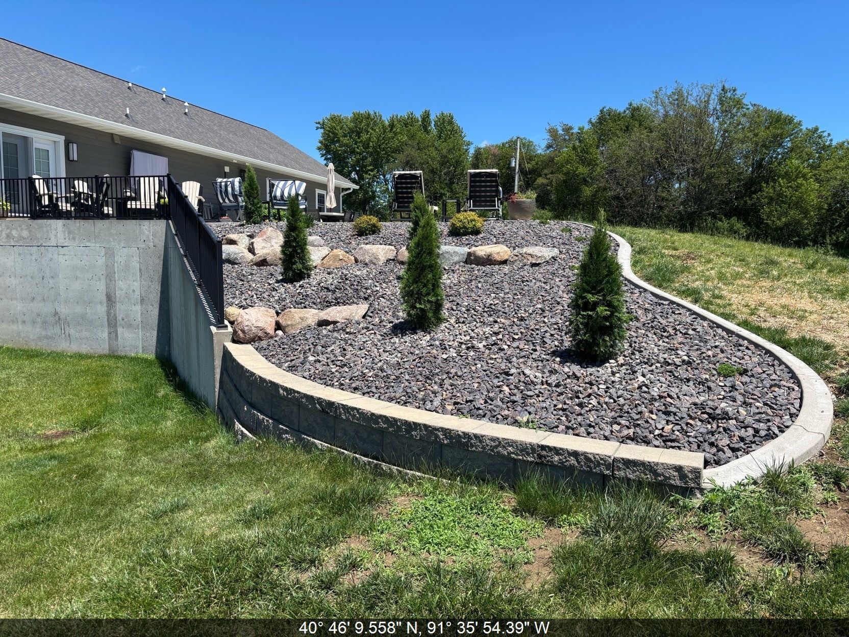 Landscaped backyard with curved stone wall, mulch beds, and small evergreen shrubs beside a house