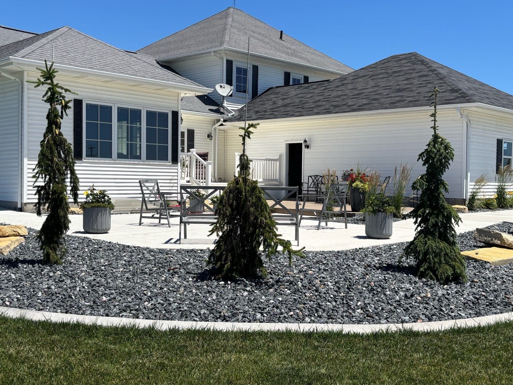 Backyard patio with white house, outdoor dining set, satellite dish, and landscaping rocks under bright sunlight