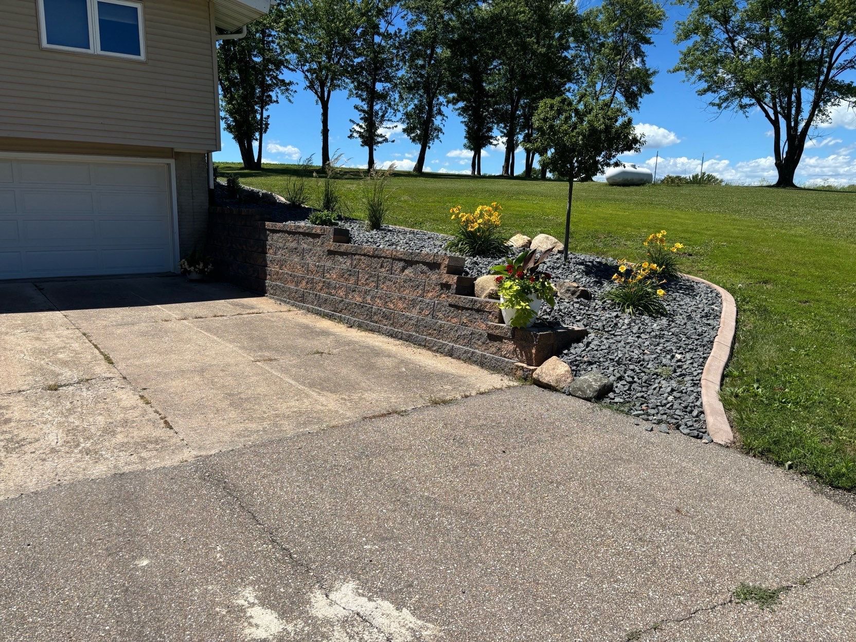 Side yard landscaping with stone retaining wall, flowers, and a curved gravel border beside a driveway and house.