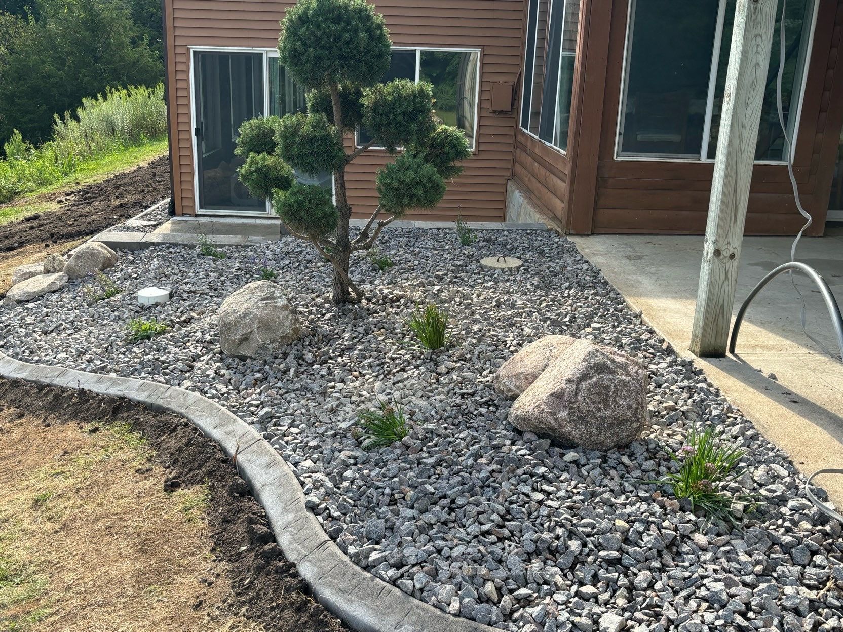 Landscaped yard with a trimmed topiary tree, decorative rocks, and a curved stone border beside a house