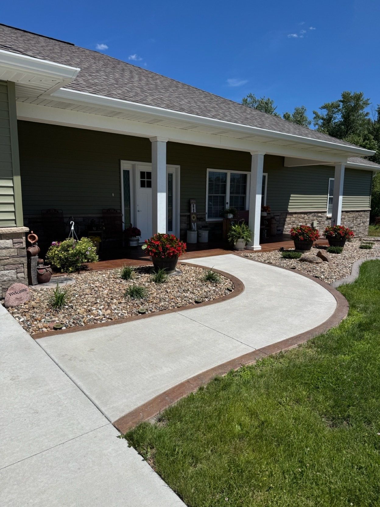 Front porch of a house with a curved concrete walkway, white columns, and landscaped flower beds under a blue sky