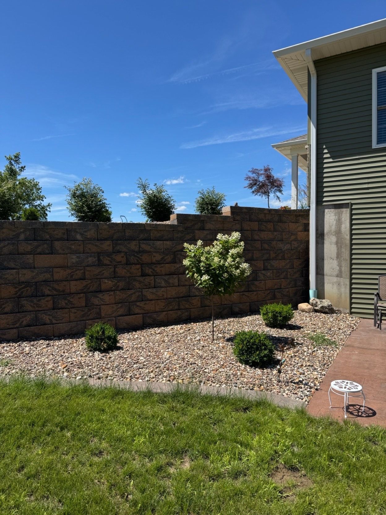 Backyard with stone retaining wall, gravel bed, shrubs, and a house exterior under a clear blue sky