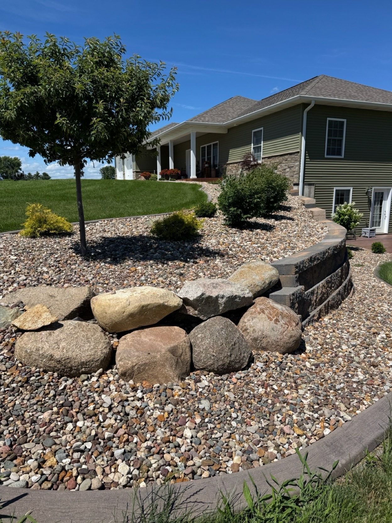 Landscaped front yard with rock garden, gravel, shrubs, and a tree beside a house