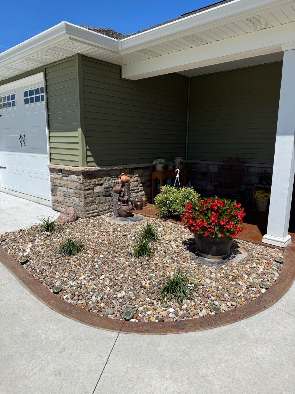 Front porch with olive siding, white columns, rock garden, and red flowers beside a garage door