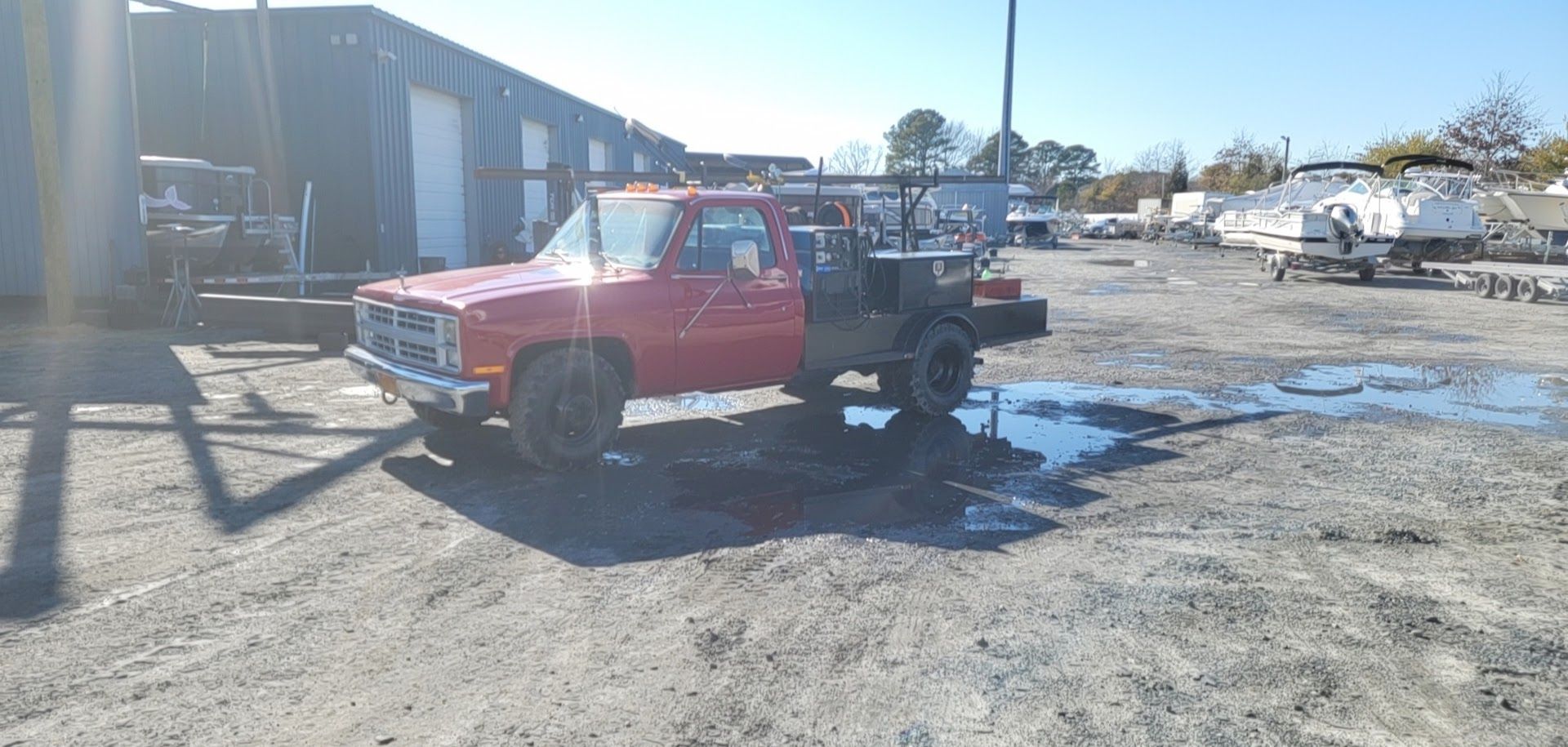 Red pickup truck with a flatbed parked in a muddy lot. A building is behind it on a sunny day.