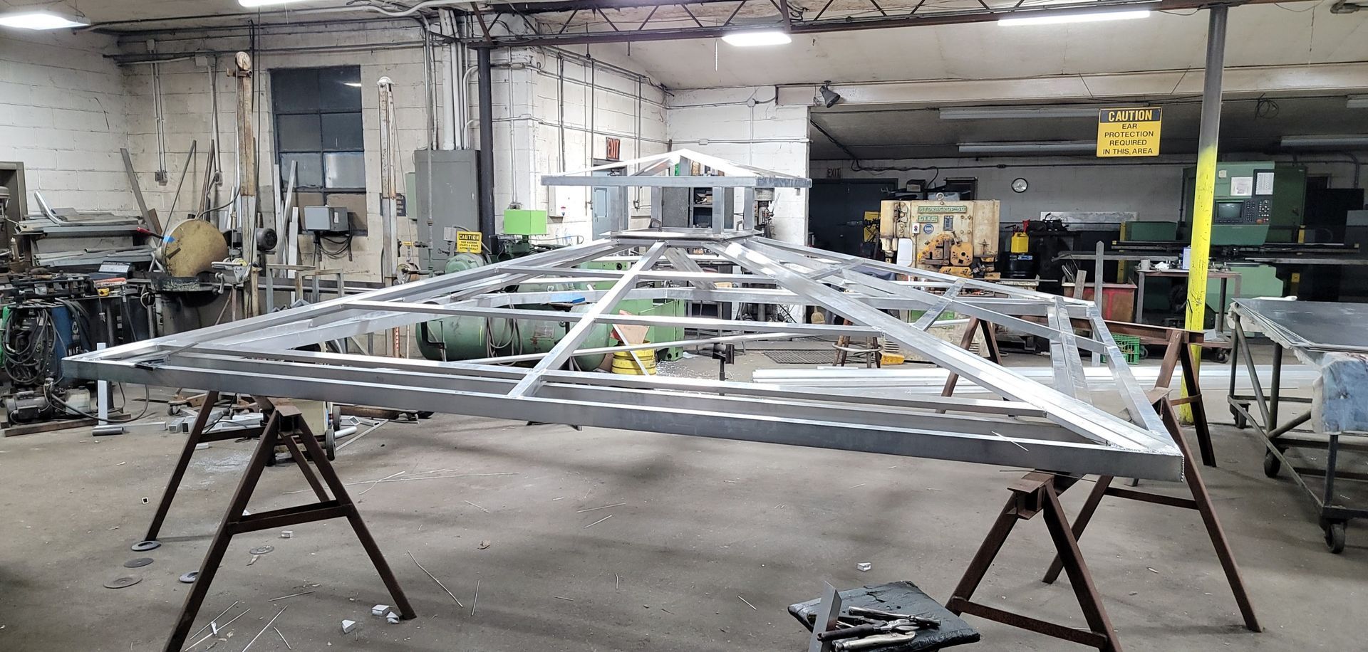Metal frame structure, possibly a gazebo, under construction in a workshop, on sawhorses.