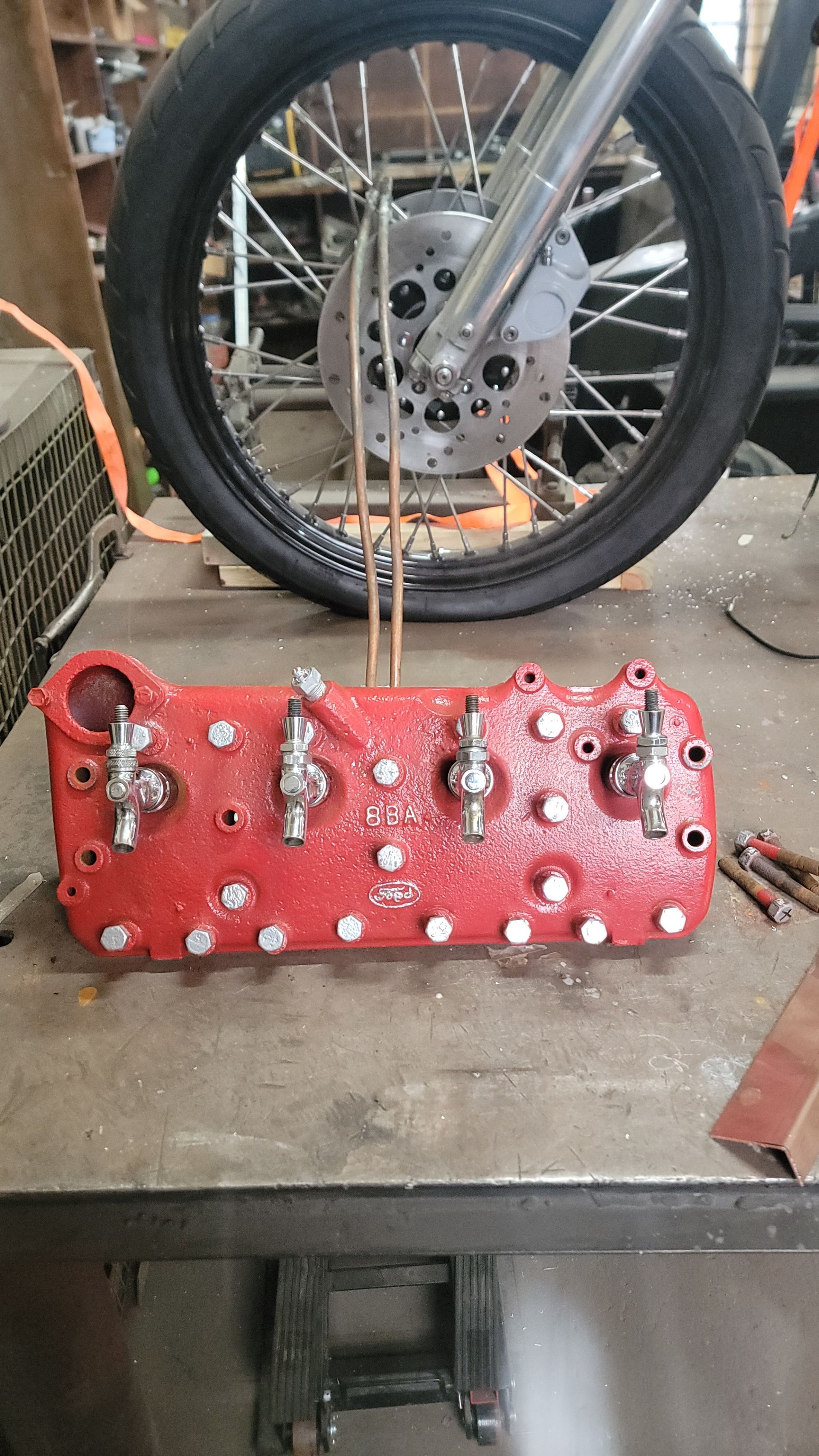 Red cylinder head with valves, sitting on a metal workbench. Motorcycle wheel in the background.