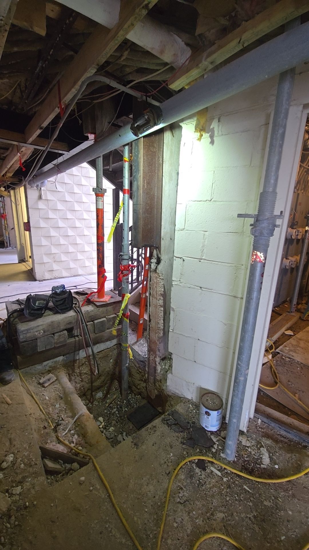 Basement construction site with support jacks, pipes, exposed wall, and excavated area.