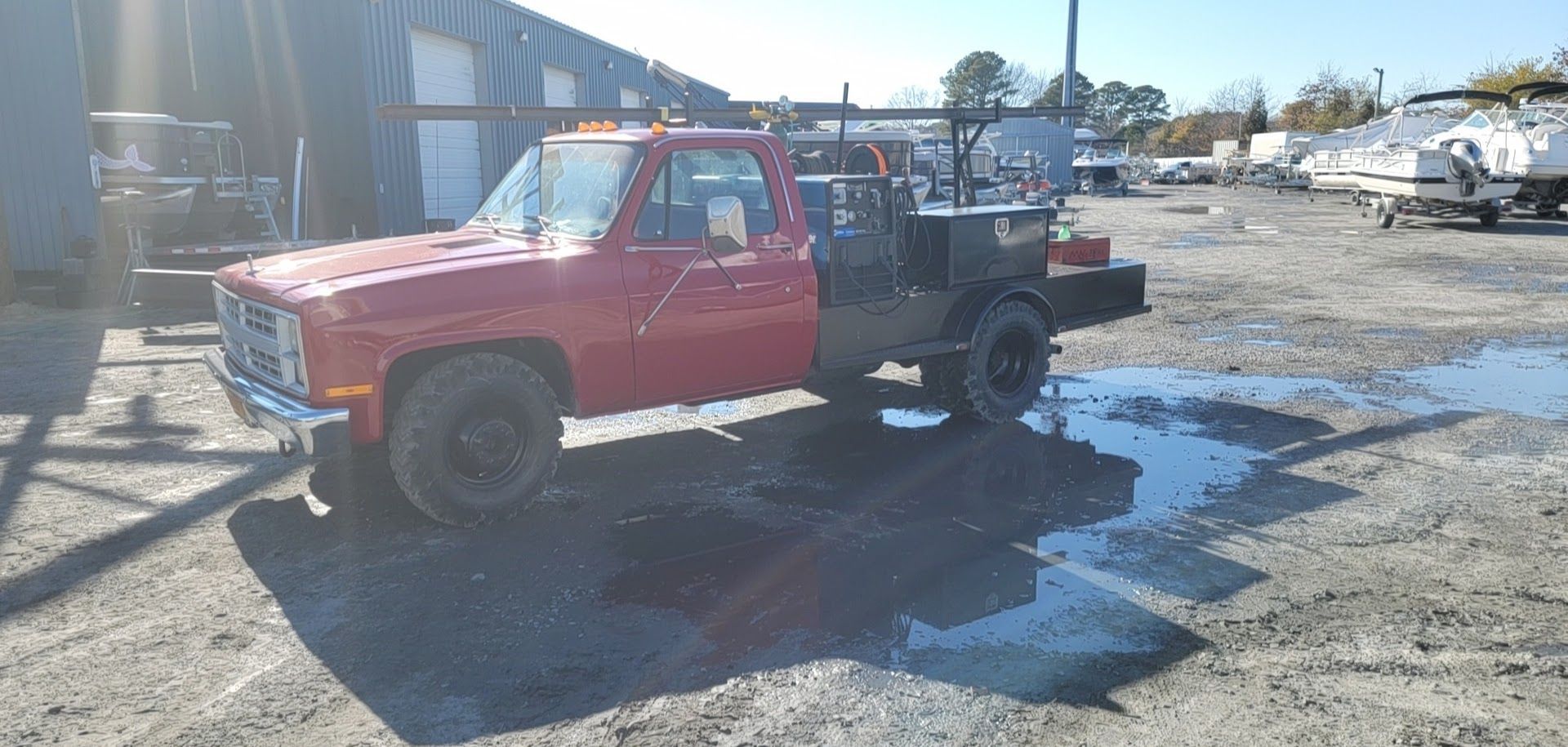 Red pickup truck with a utility bed parked in a gravel lot on a sunny day.