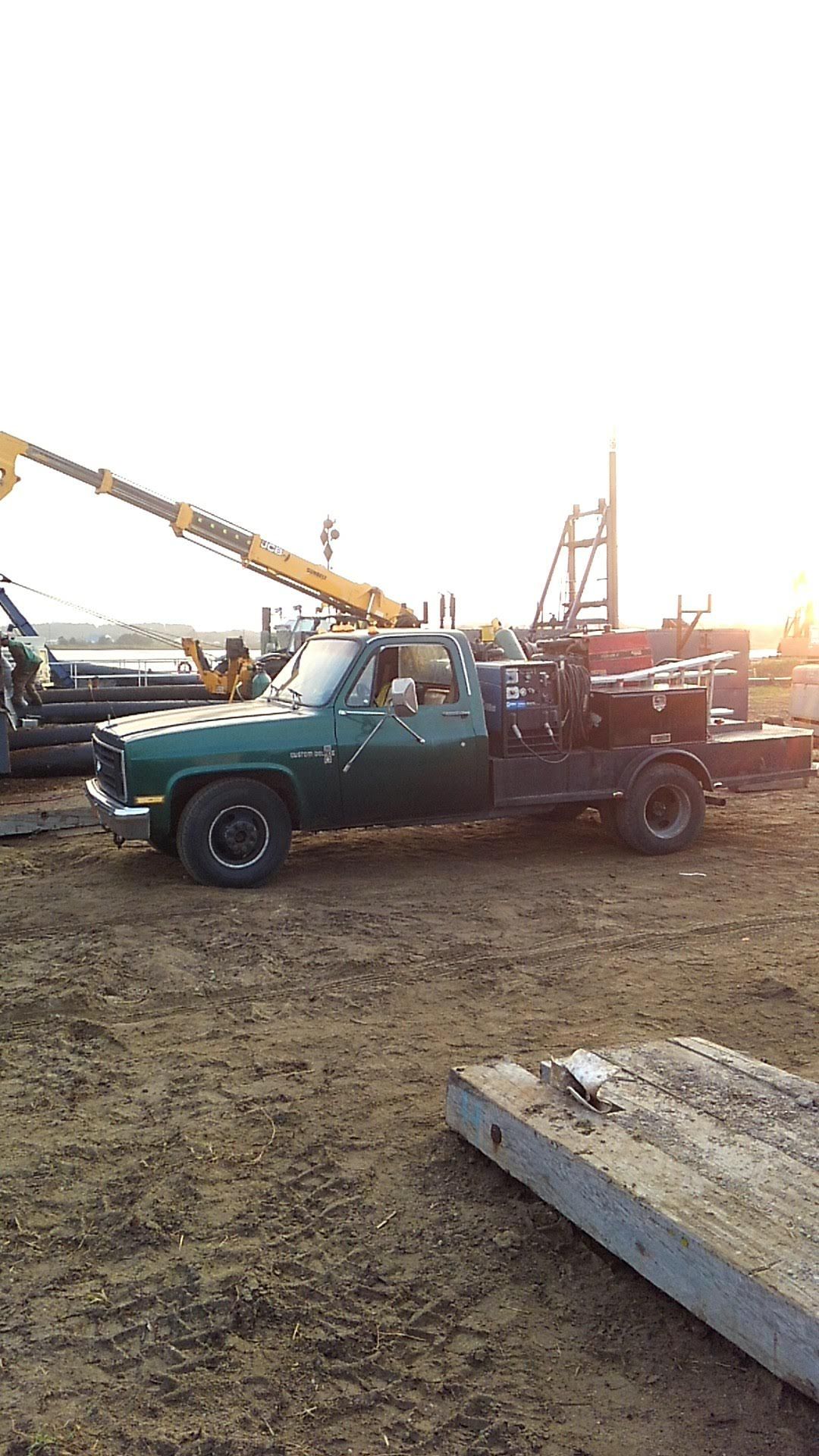 Green work truck with tools parked on dirt. A crane and equipment are in the background, likely at a worksite.