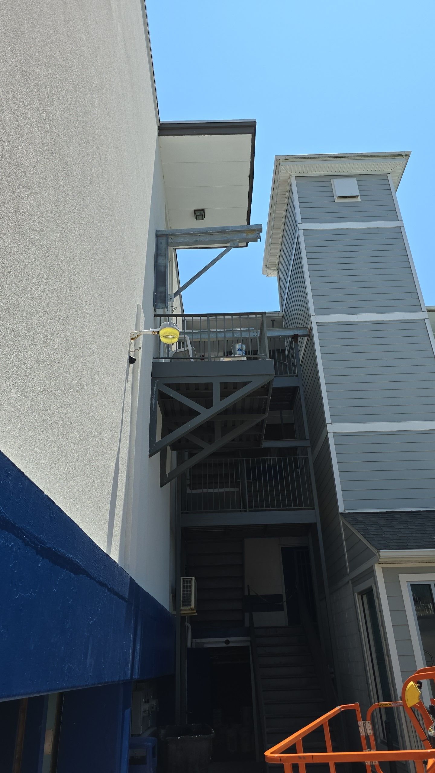 Exterior stairwell between two buildings. Grey stairs and wooden support structure. Blue sky.