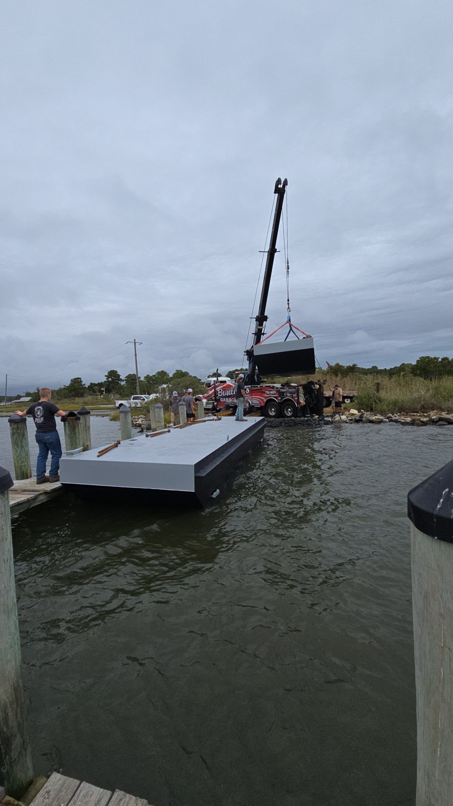 Crane lifting a dark structure onto a floating dock in a waterway. Cloudy sky, man observing.