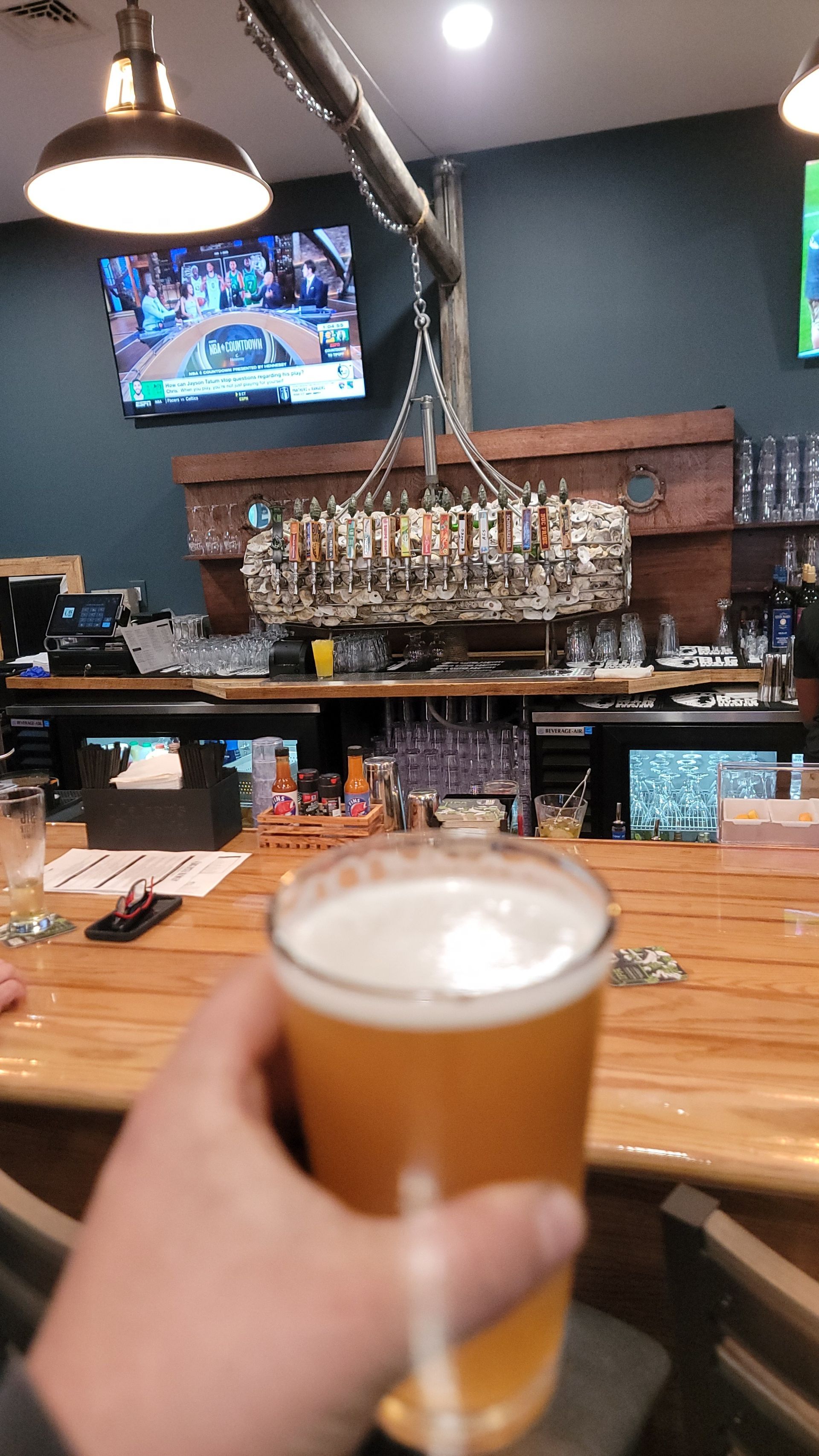 Person holding a pint of beer at a bar. Wooden bar, overhead lighting, TVs playing sports.