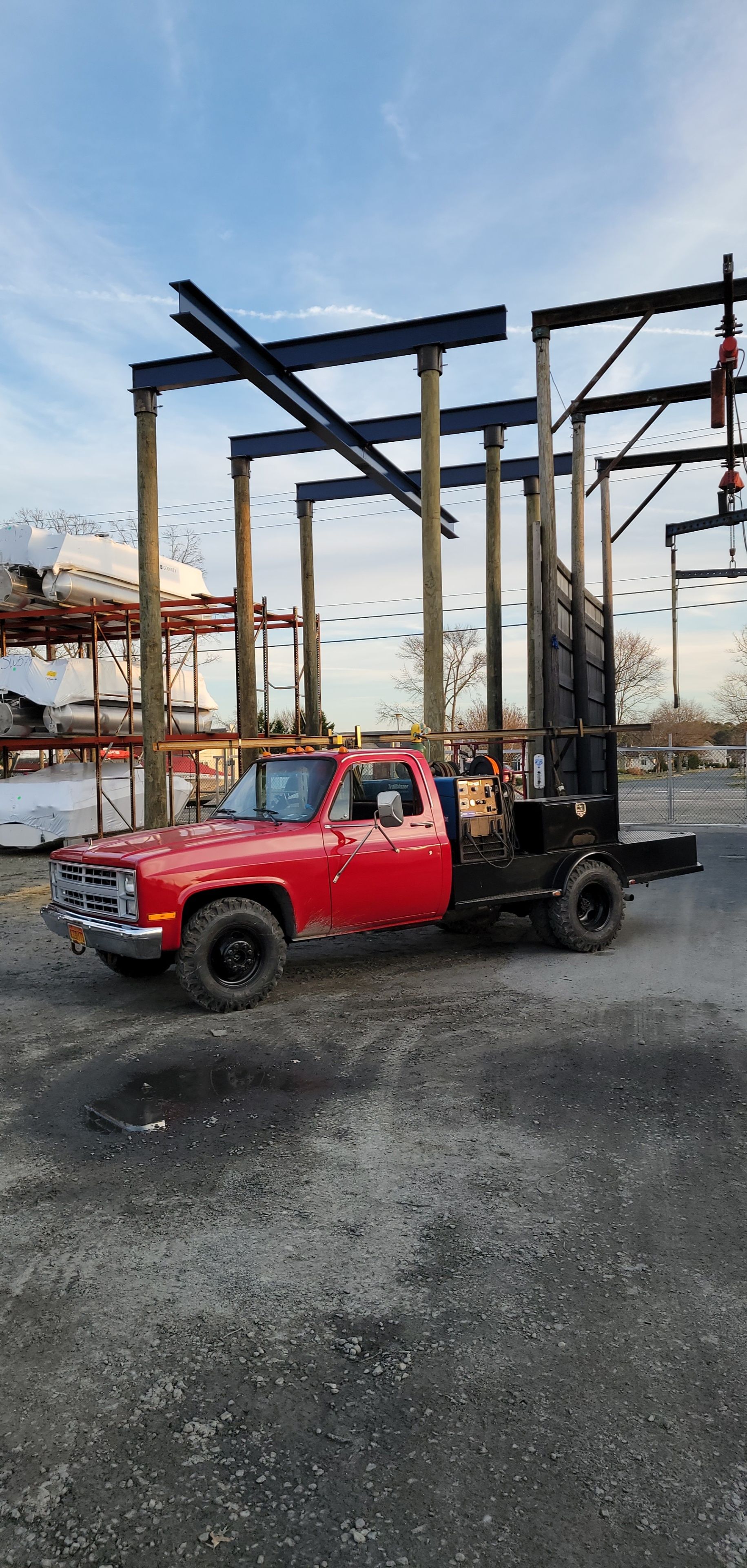 Red pickup truck with a flatbed in front of steel structure under a blue sky.