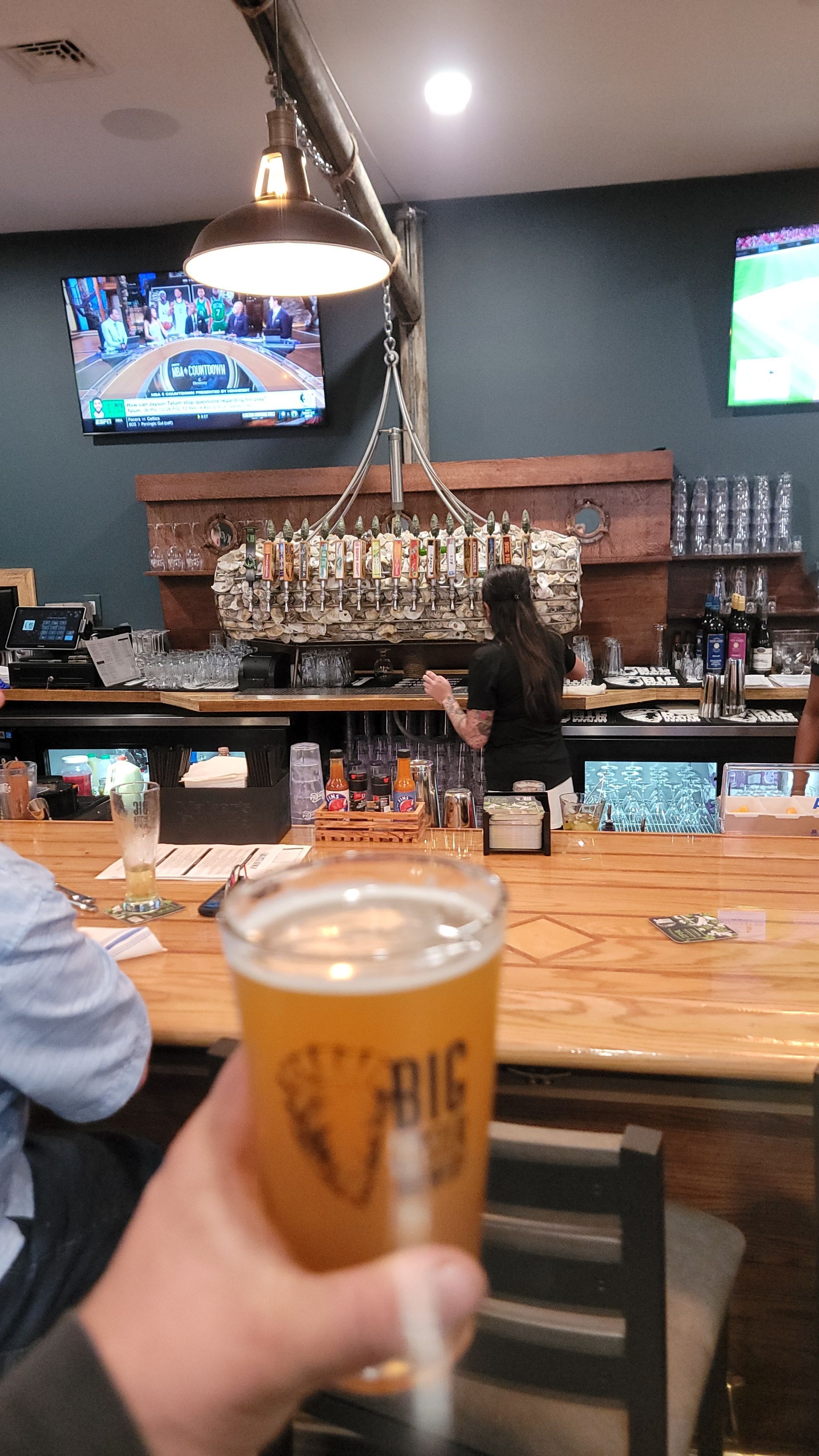A hand holding a beer at a bar. A bartender works behind the bar with bottles hanging above. A TV plays a game.