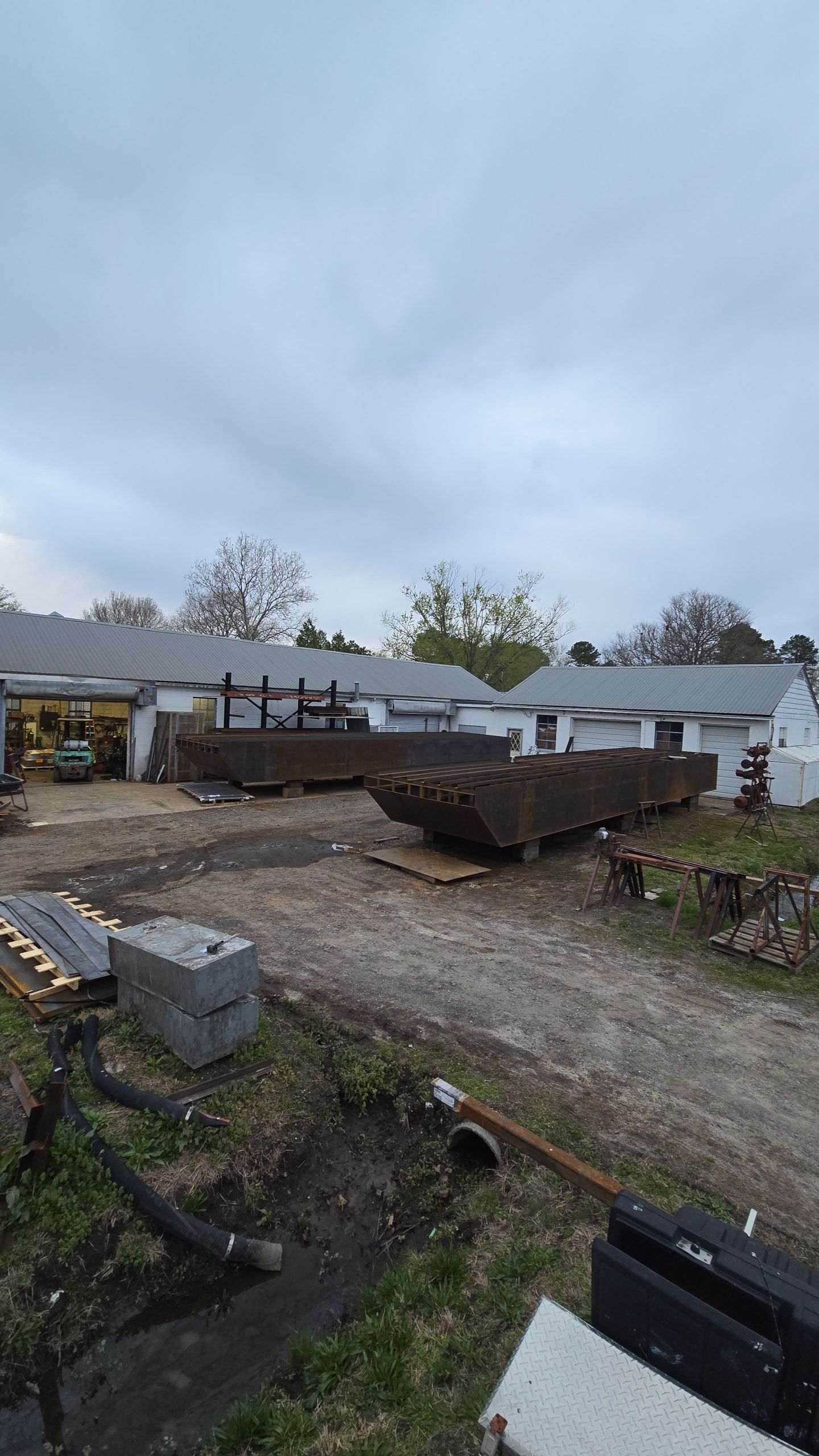 Dirt lot with lumber and concrete blocks in front of white buildings under overcast sky.