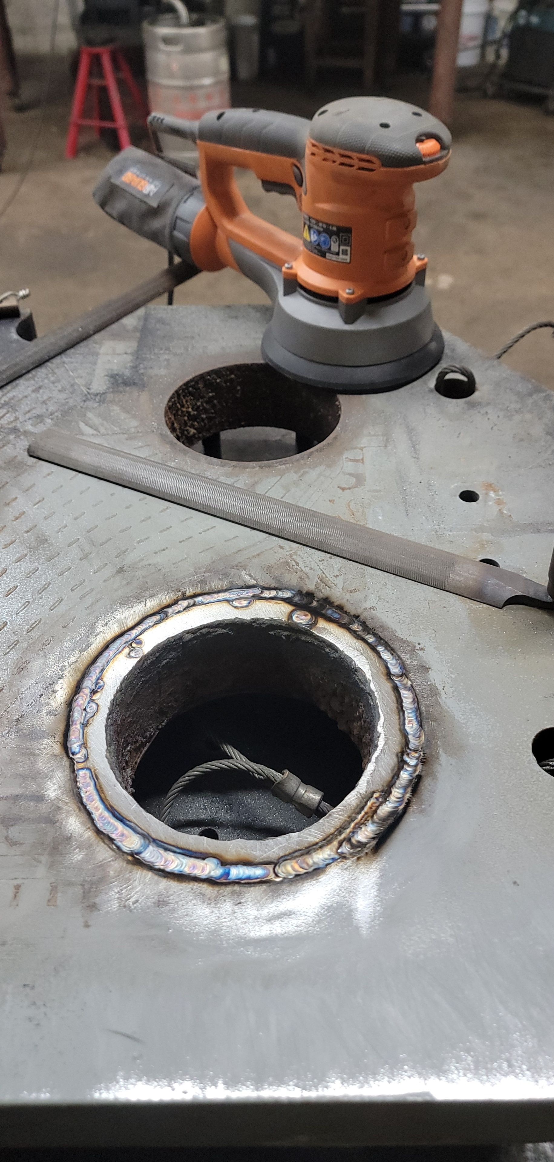 An orange sander rests on a metal surface with two holes. A file lies across the surface.