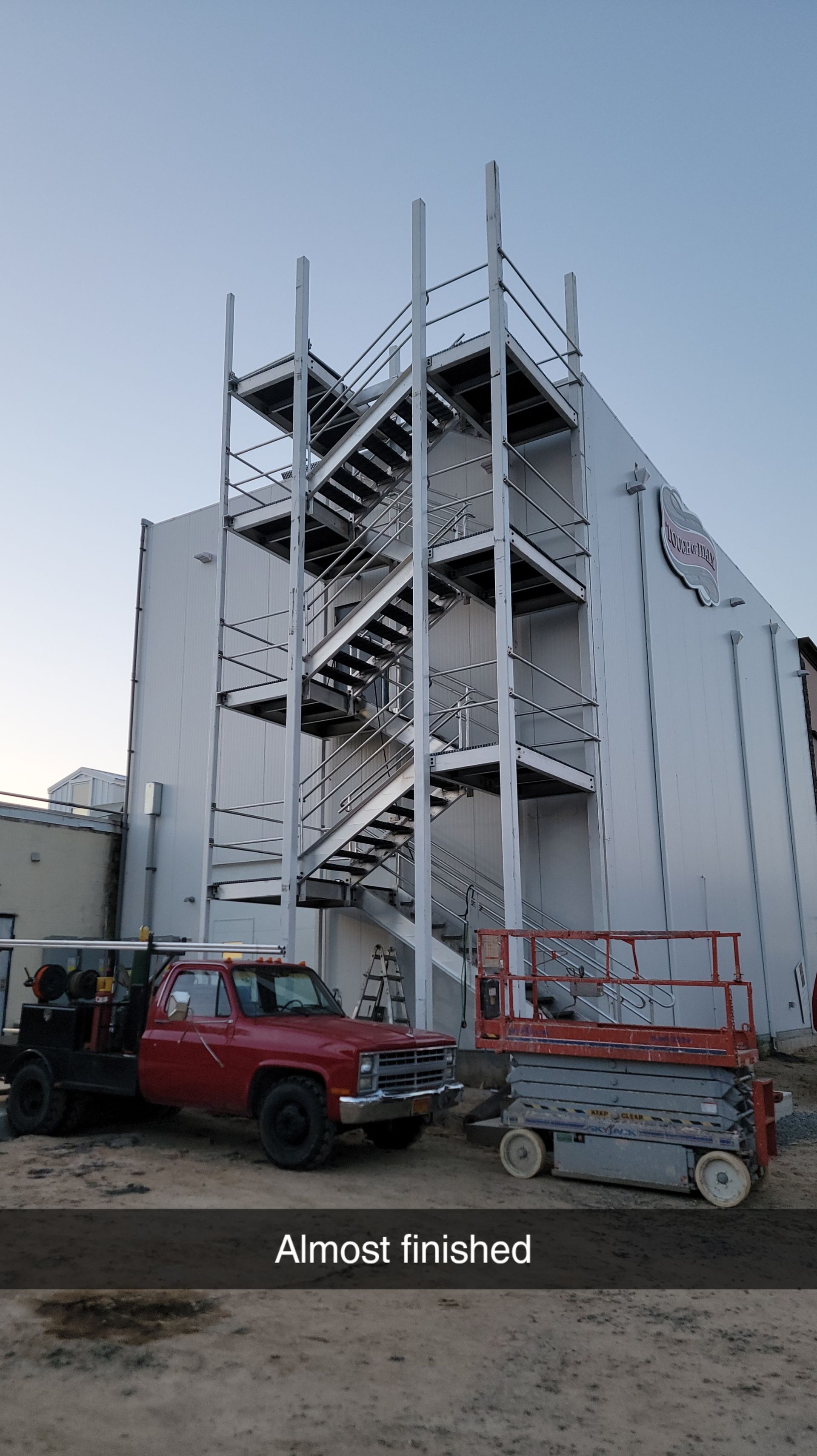 Exterior metal staircase attached to a white building; a red truck and lift sit below. Text reads: 