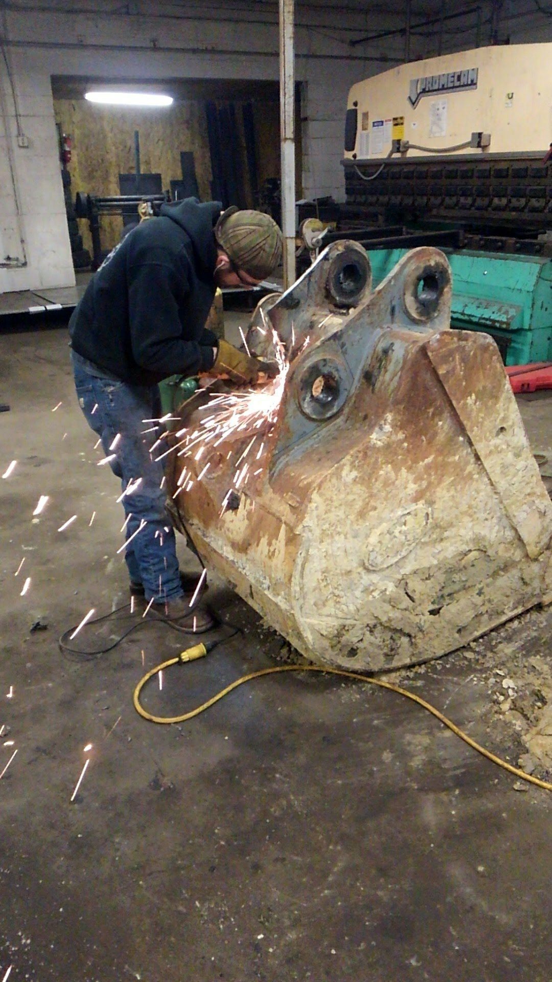Person grinding metal on an excavator bucket, creating sparks, in a workshop.