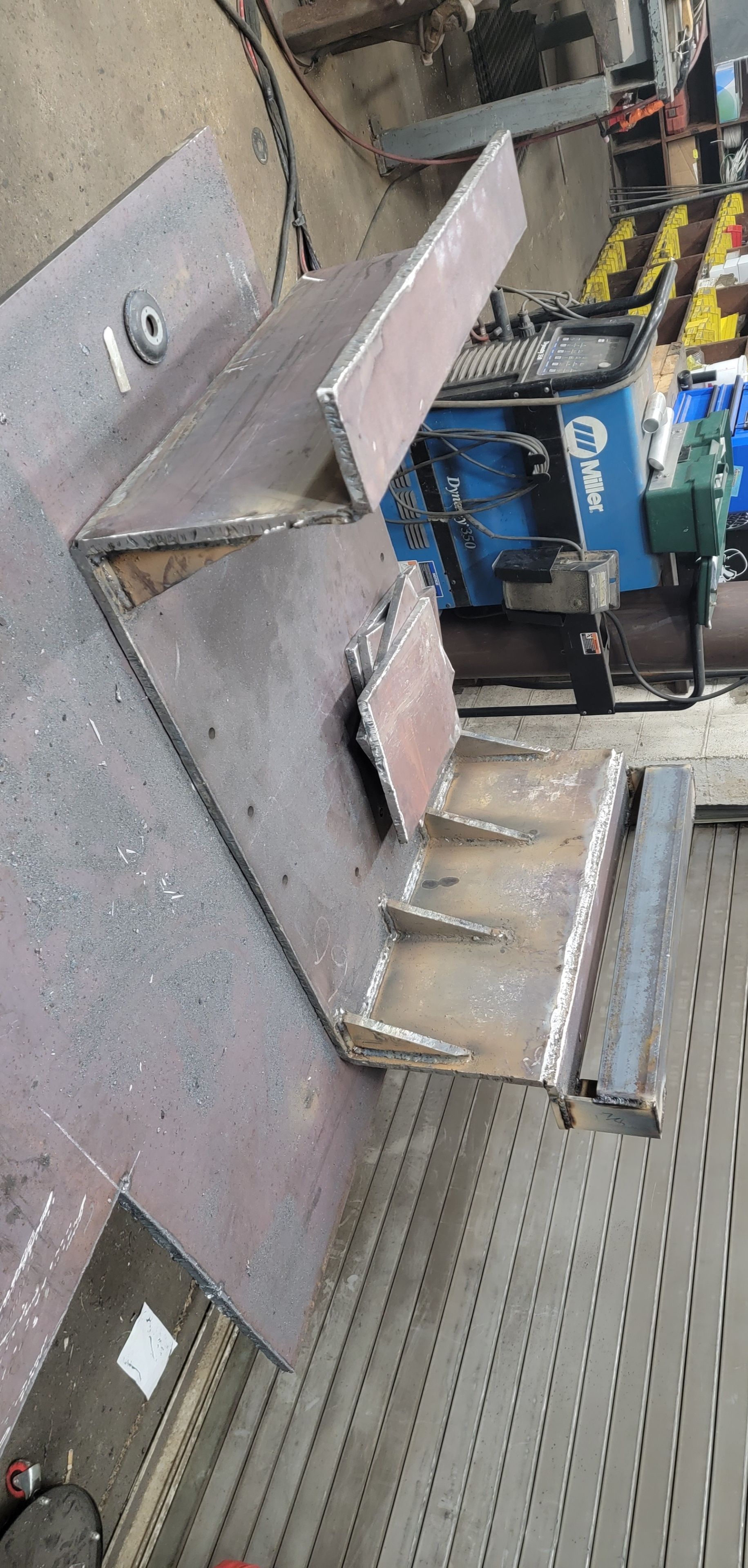 Welded steel fabrication, possibly a bracket or support, on a metal workbench. Miller welding machine in background.