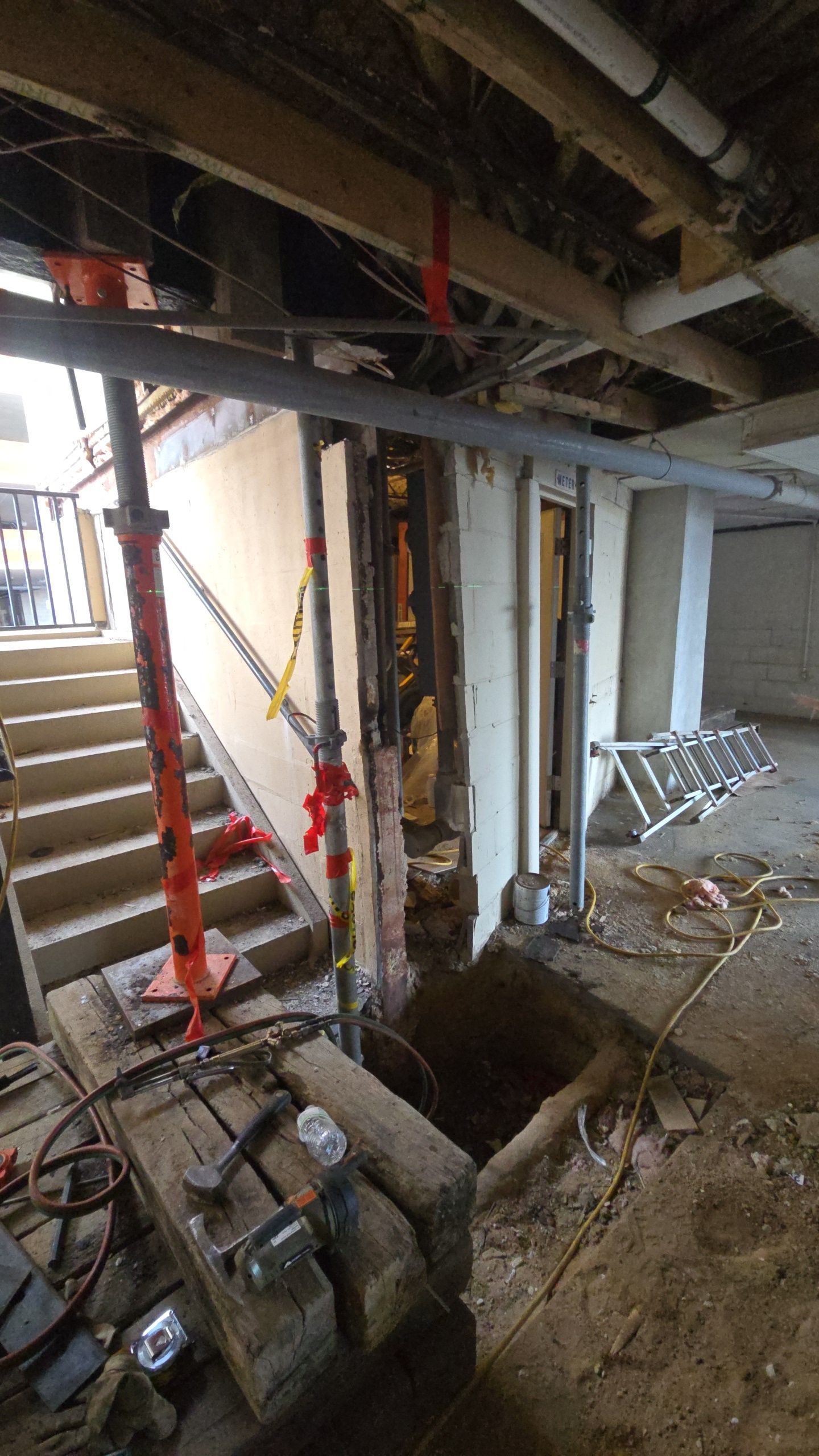 Basement under construction, with exposed framing, stairs, support jacks, and a ladder.