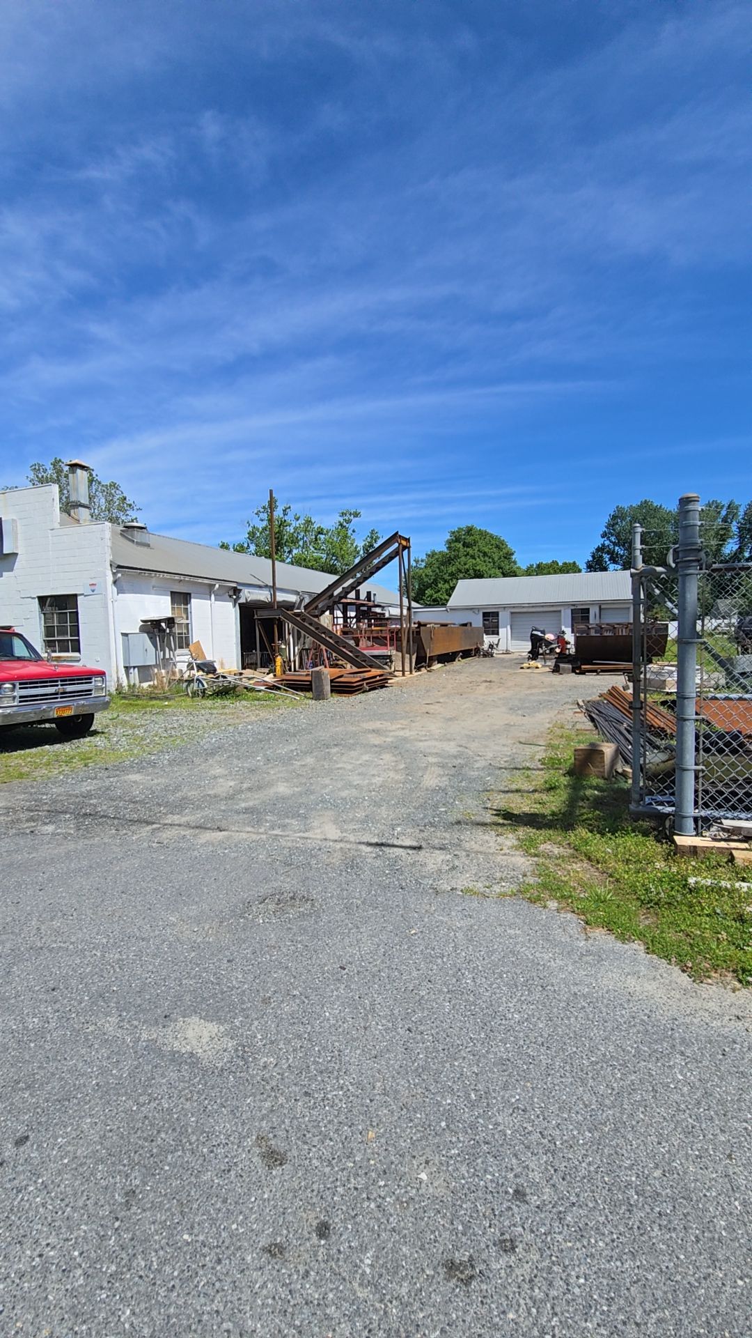 Gravel parking area with a white building, wooden structure, and red truck under a bright blue sky.