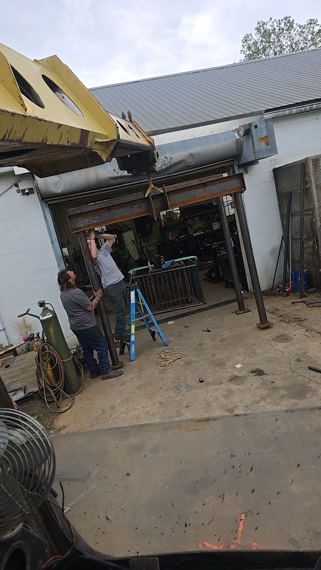 Two people working on metal structure by a building; one on a ladder, a large yellow object overhead.