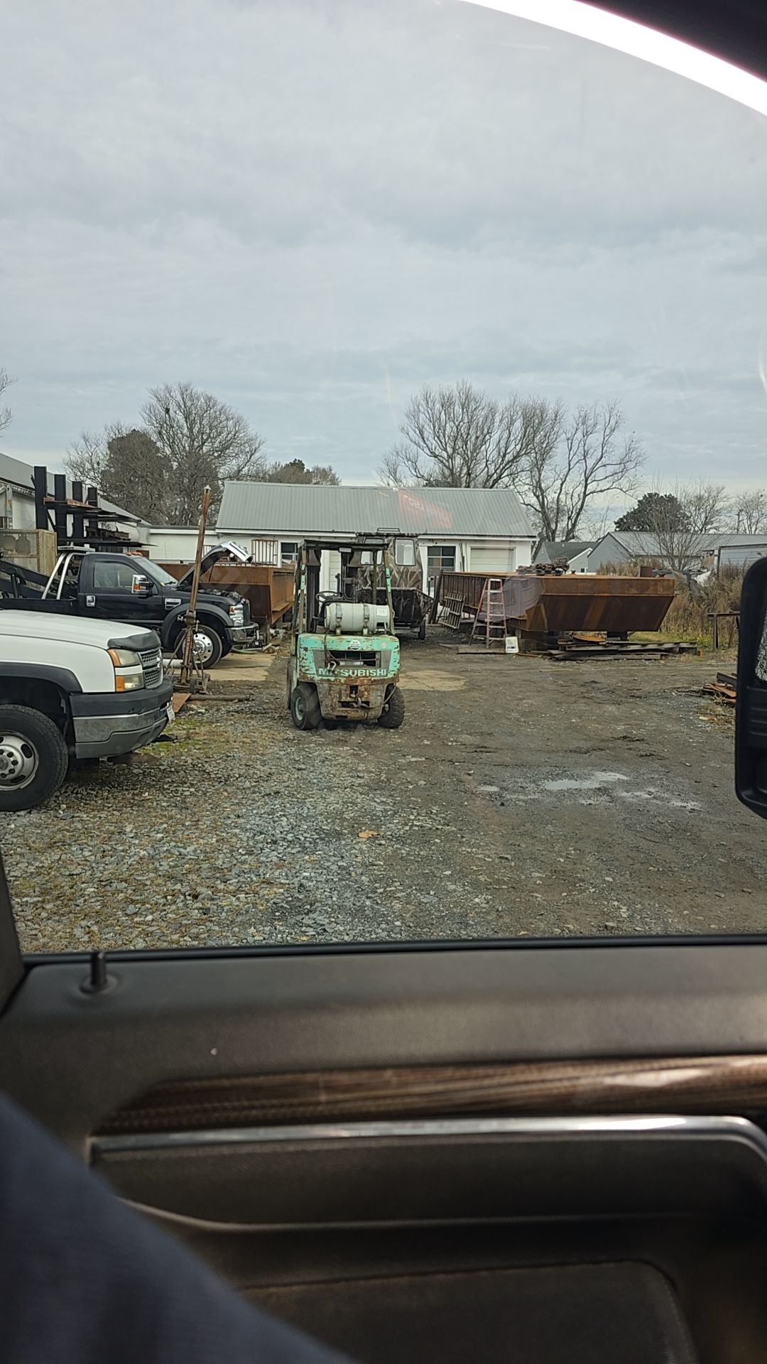A gravel lot at a salvage yard with trucks, a skid steer, and metal scraps. Cloudy sky.