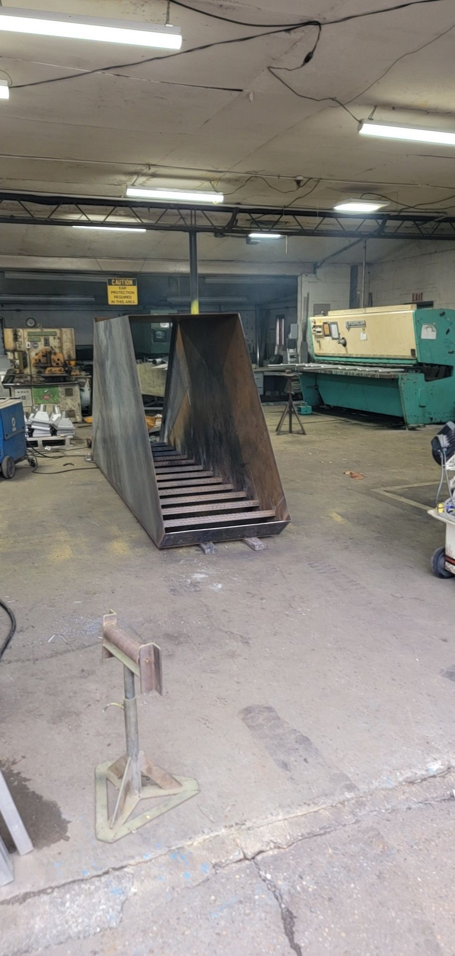 A large metal structure with a grated bottom inside a workshop. A cutting machine is in the background.