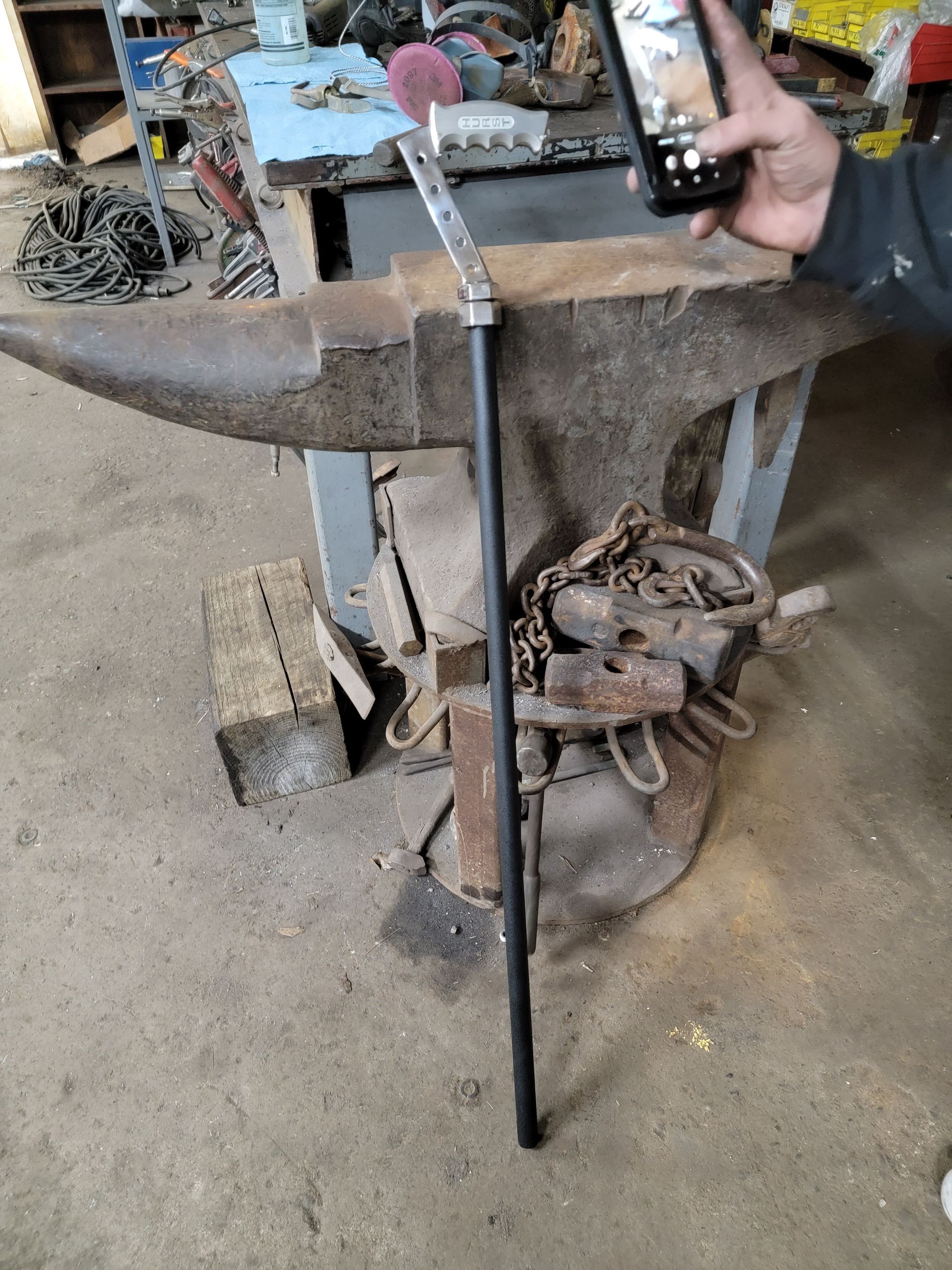 Blacksmith's workshop scene with a long metal rod, anvil, and a person taking a photo with a smartphone.
