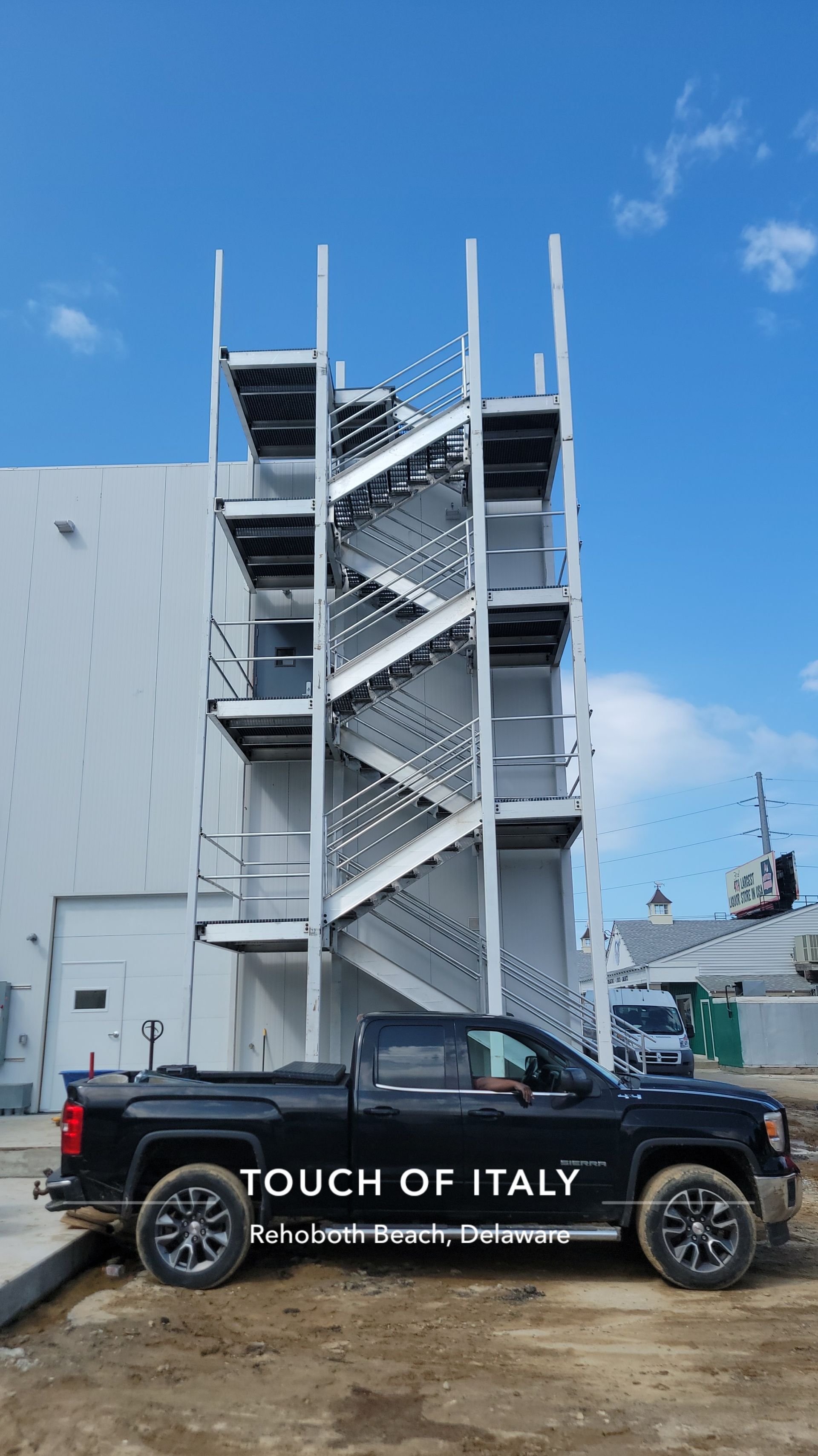 Black truck parked in front of a tall, grey metal fire escape on the side of a building. Sky in background.