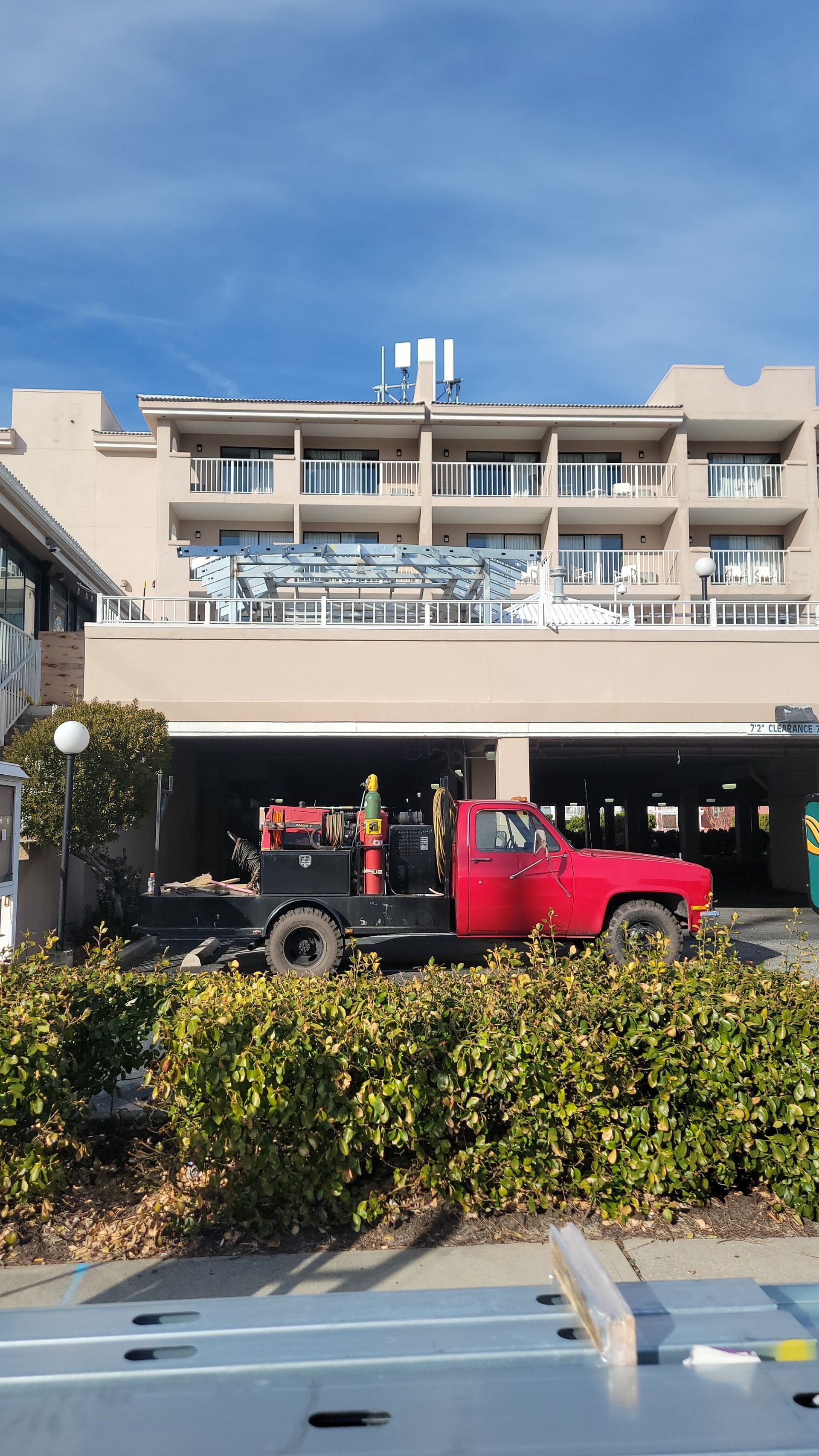 Red pickup truck parked under a building's carport. Building has balconies and a blue sky overhead.