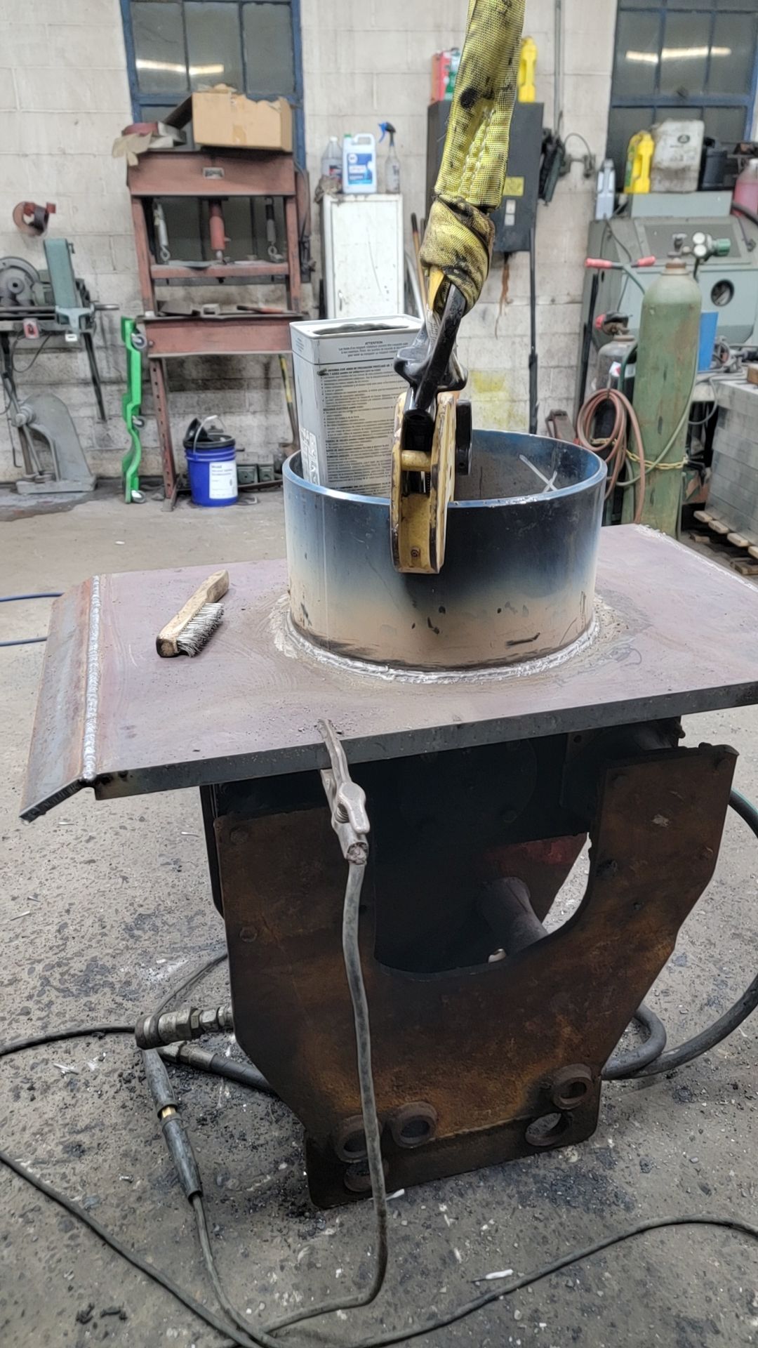A metalworker using a crane to lower a cylindrical piece into a larger steel structure inside a workshop.
