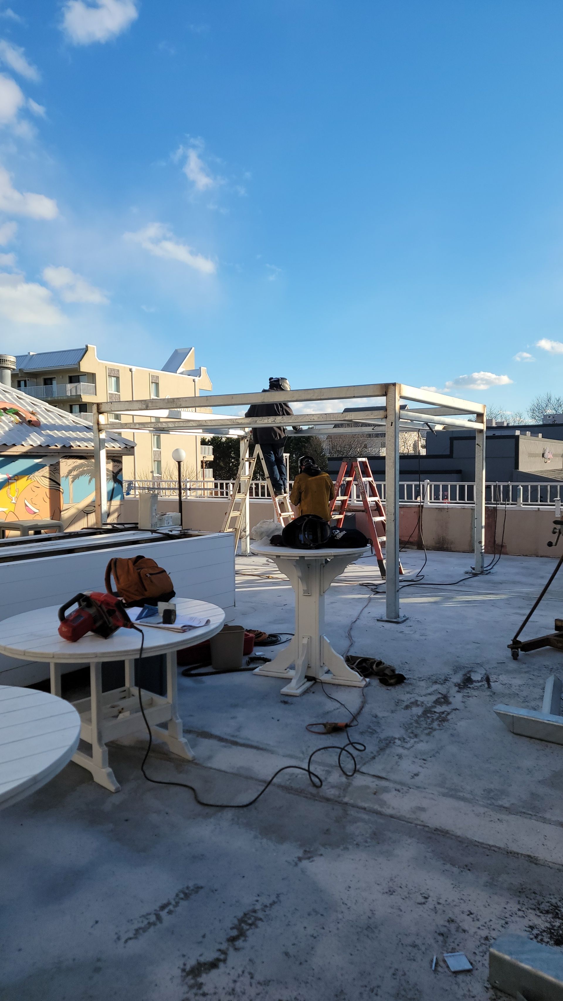 Construction on rooftop: Metal frame being built. Tools and tables are scattered on the concrete surface under a blue sky.