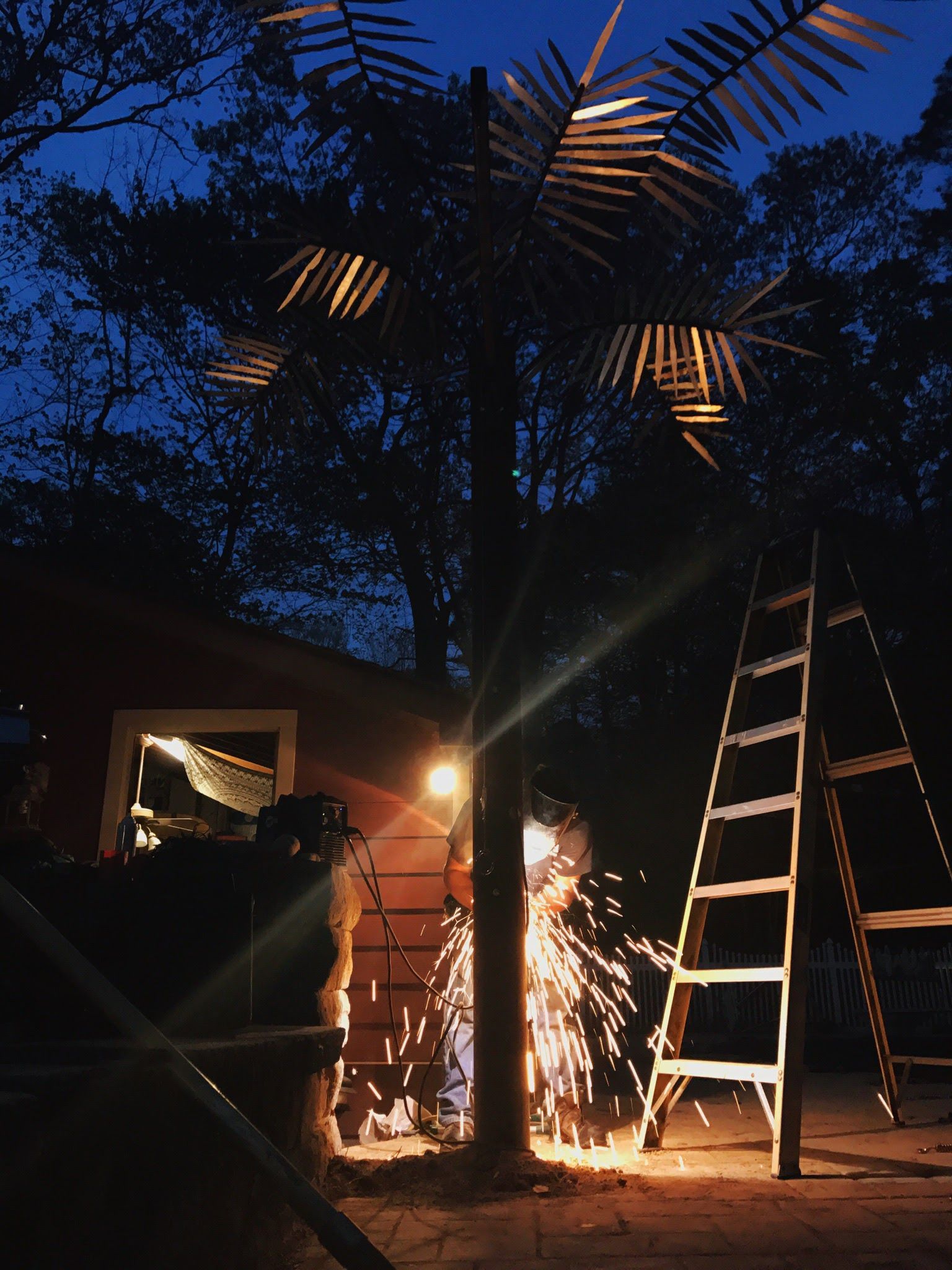 Person welding a metal palm tree at night, sparks flying. Ladder and building in the background.