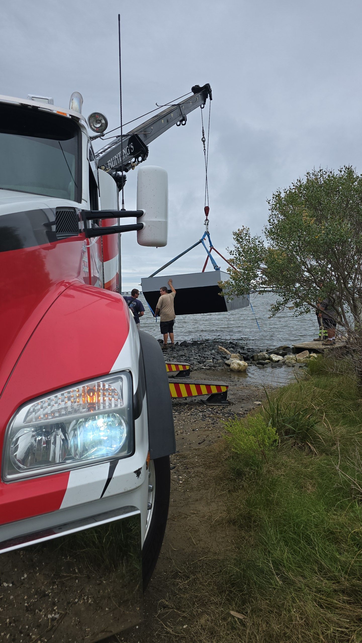 A crane lifting a large, dark rectangular object near a body of water. A red and white truck is in the foreground.