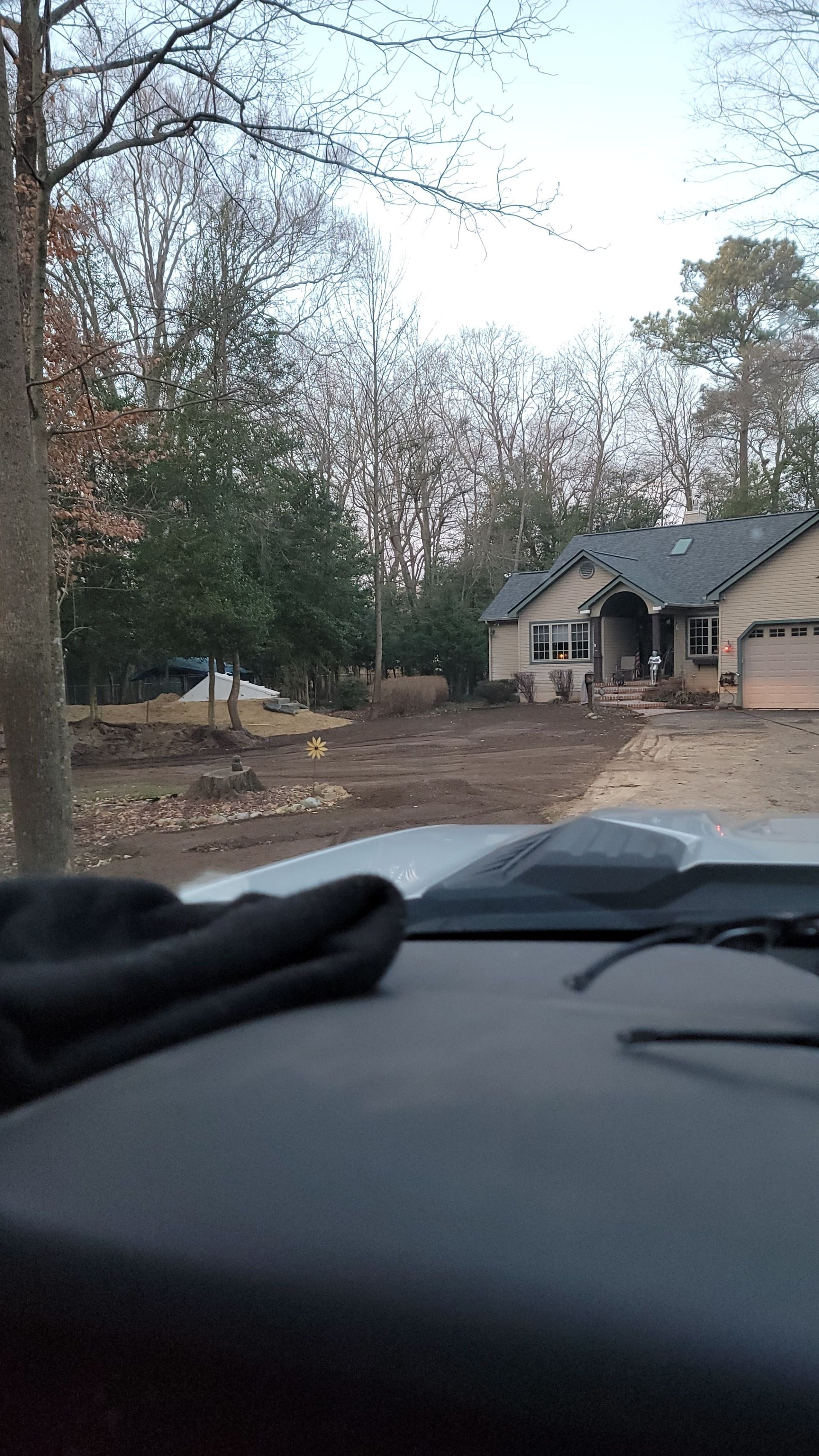 House with bare yard, viewed from inside vehicle. Trees, overcast sky.
