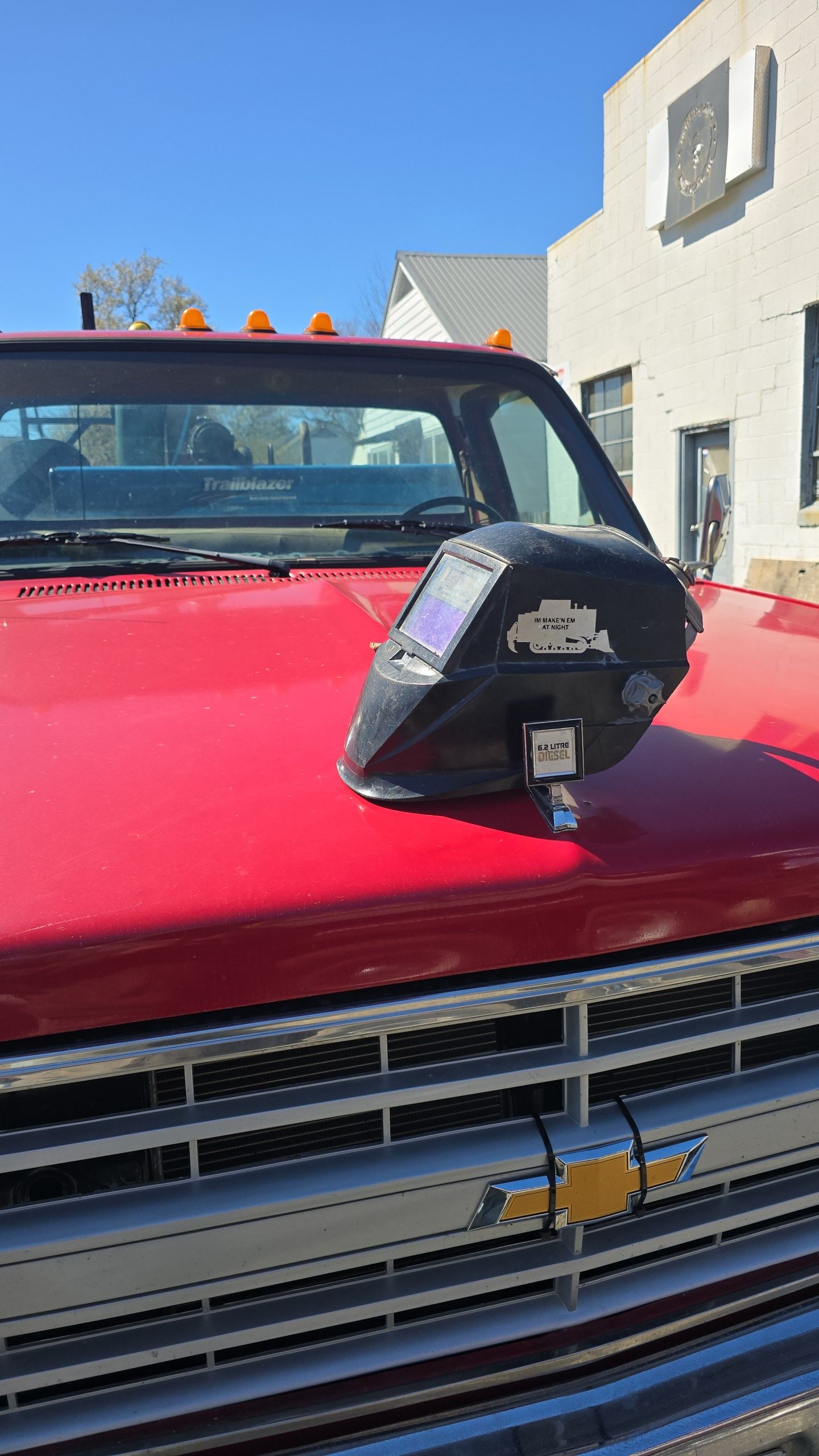 Red truck hood with a black welding helmet resting on it, in front of a white building.