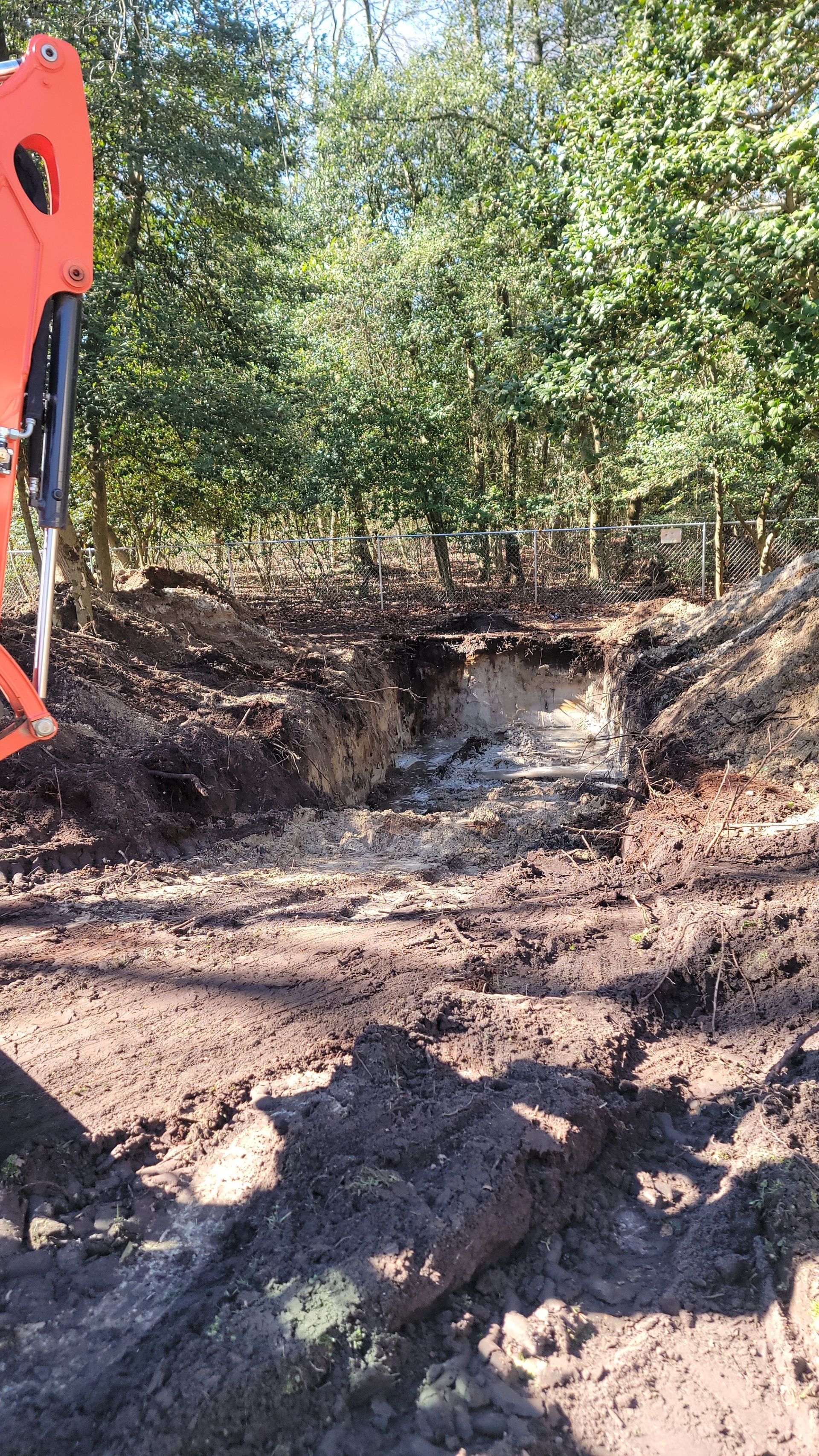 An excavator arm over a trench in the woods. The trench is dug in dirt and clay.