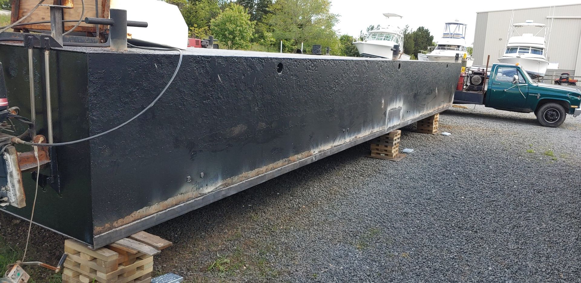 Long, black, rectangular steel structure on wooden supports; green truck in gravel lot.