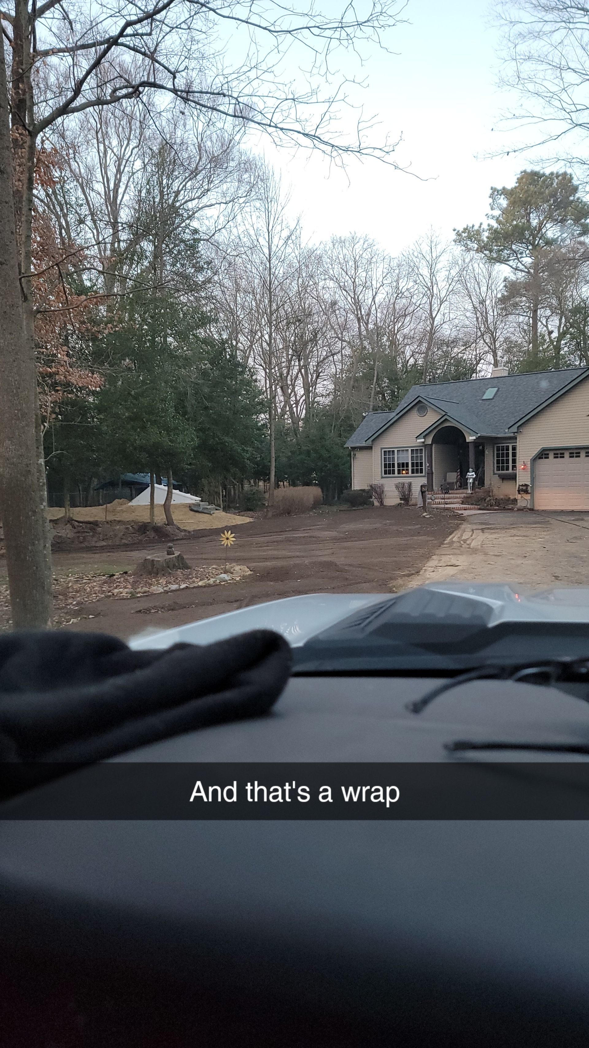 View from a vehicle's dashboard of a house with a dirt yard and bare trees. Text: 