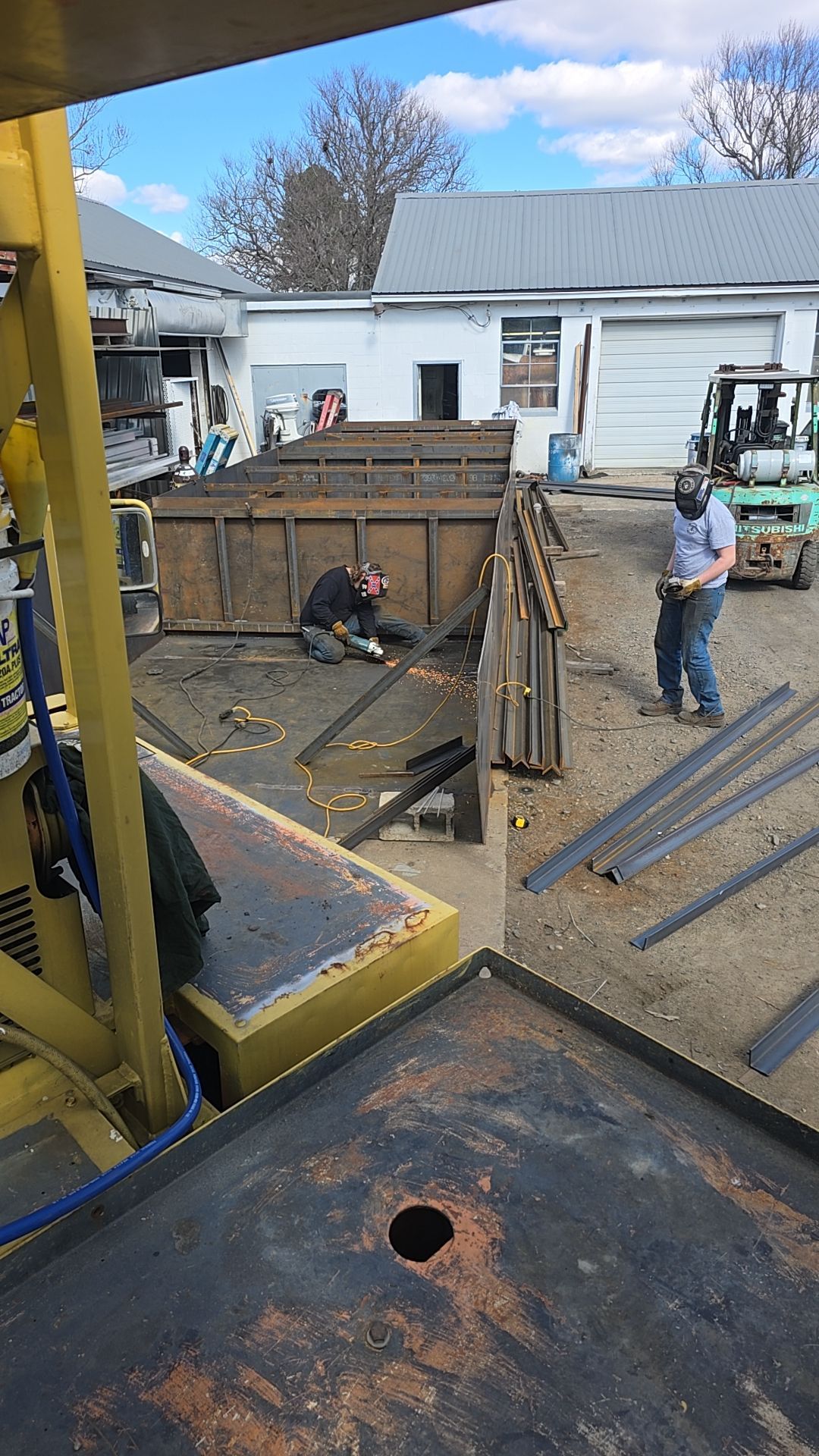 Two workers welding metal in an outdoor industrial area, using sparks, with a large container and metal pieces.