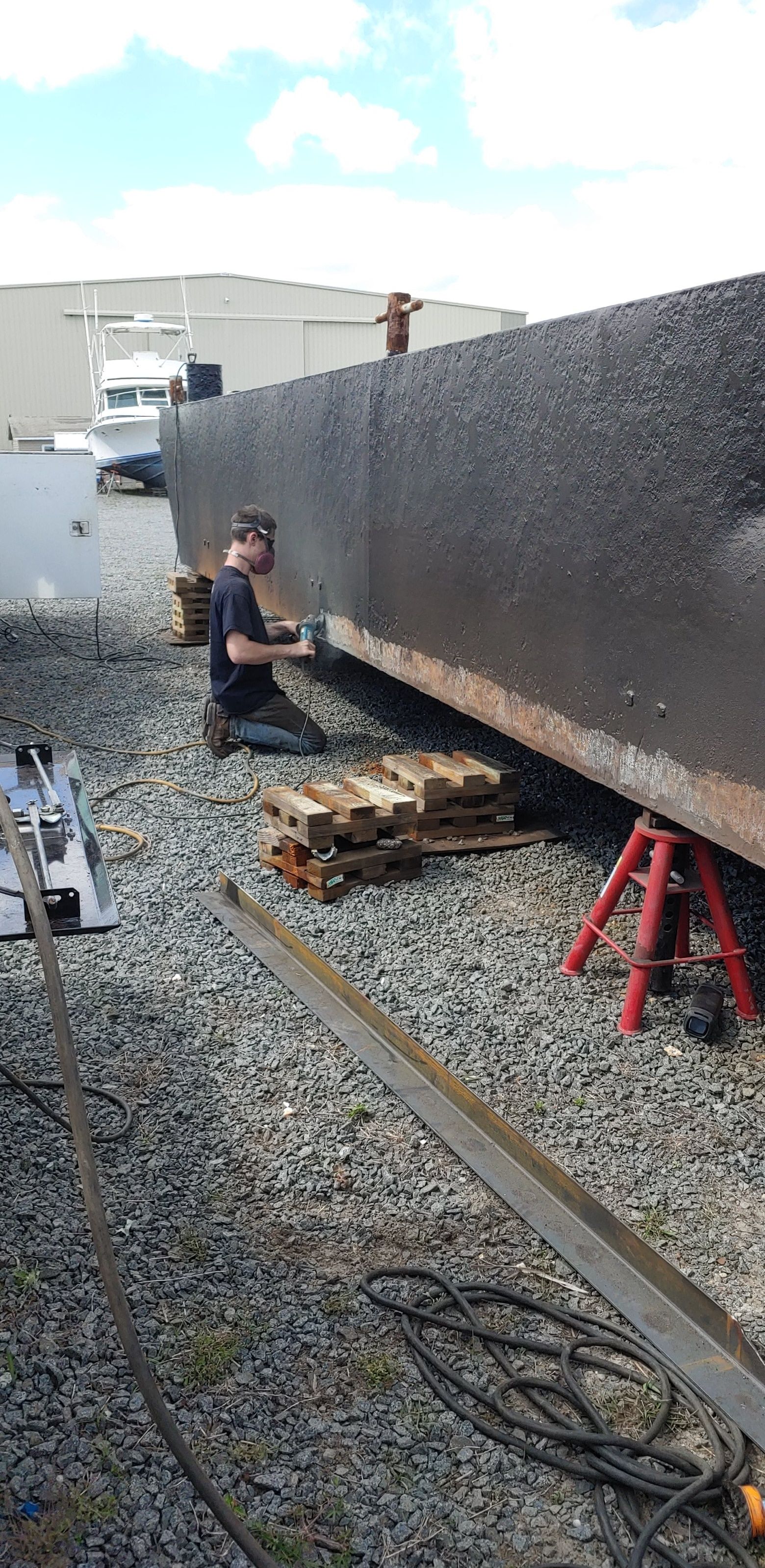 A person works on a large, dark rectangular structure. It is supported by jacks and pallets on gravel.