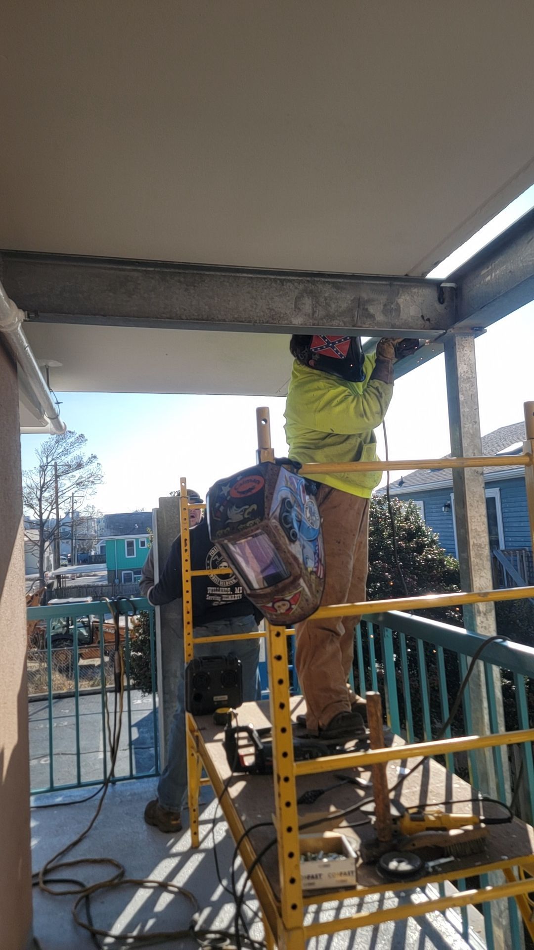 Two welders working on a metal beam, on scaffolding, outdoors. One welds, one assists.