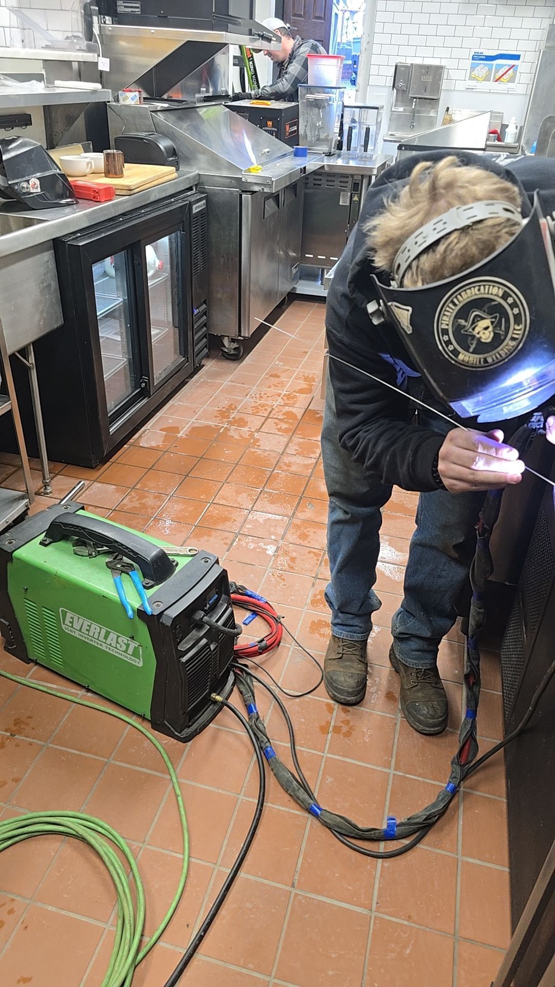 Person welding near a green welding machine inside a commercial kitchen; sparks, visible welding mask.