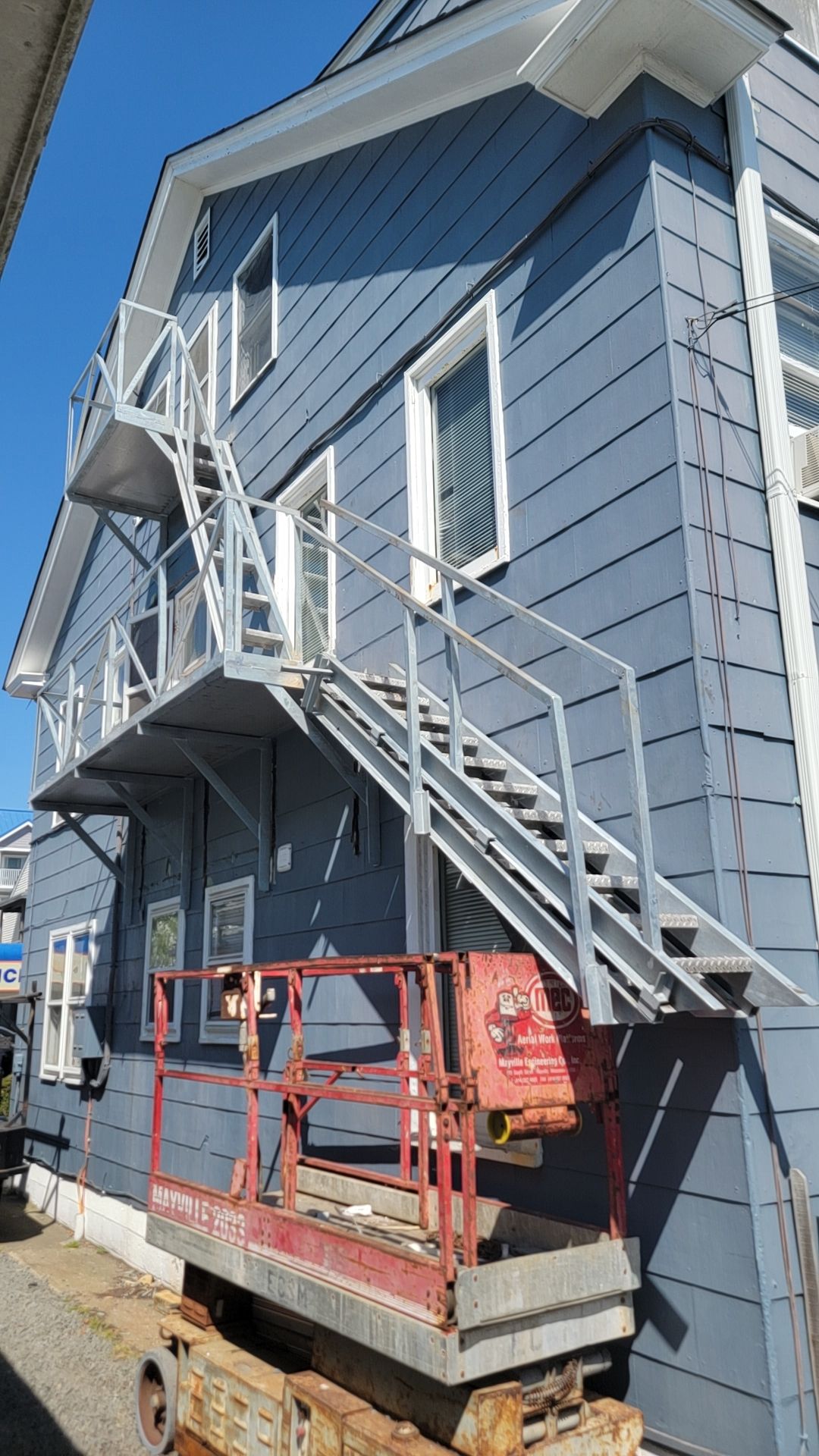 Exterior view of a three-story building with a metal fire escape. A lift is beneath the stairs. Blue siding.