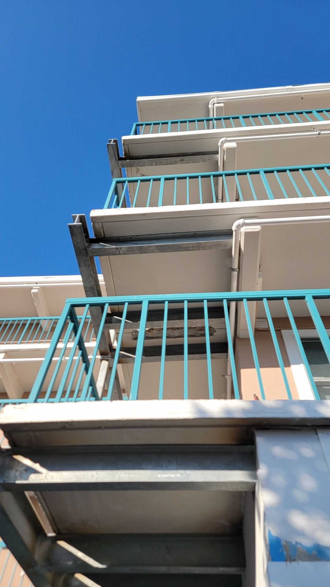 Multi-story building with turquoise railings and steel supports against a blue sky.