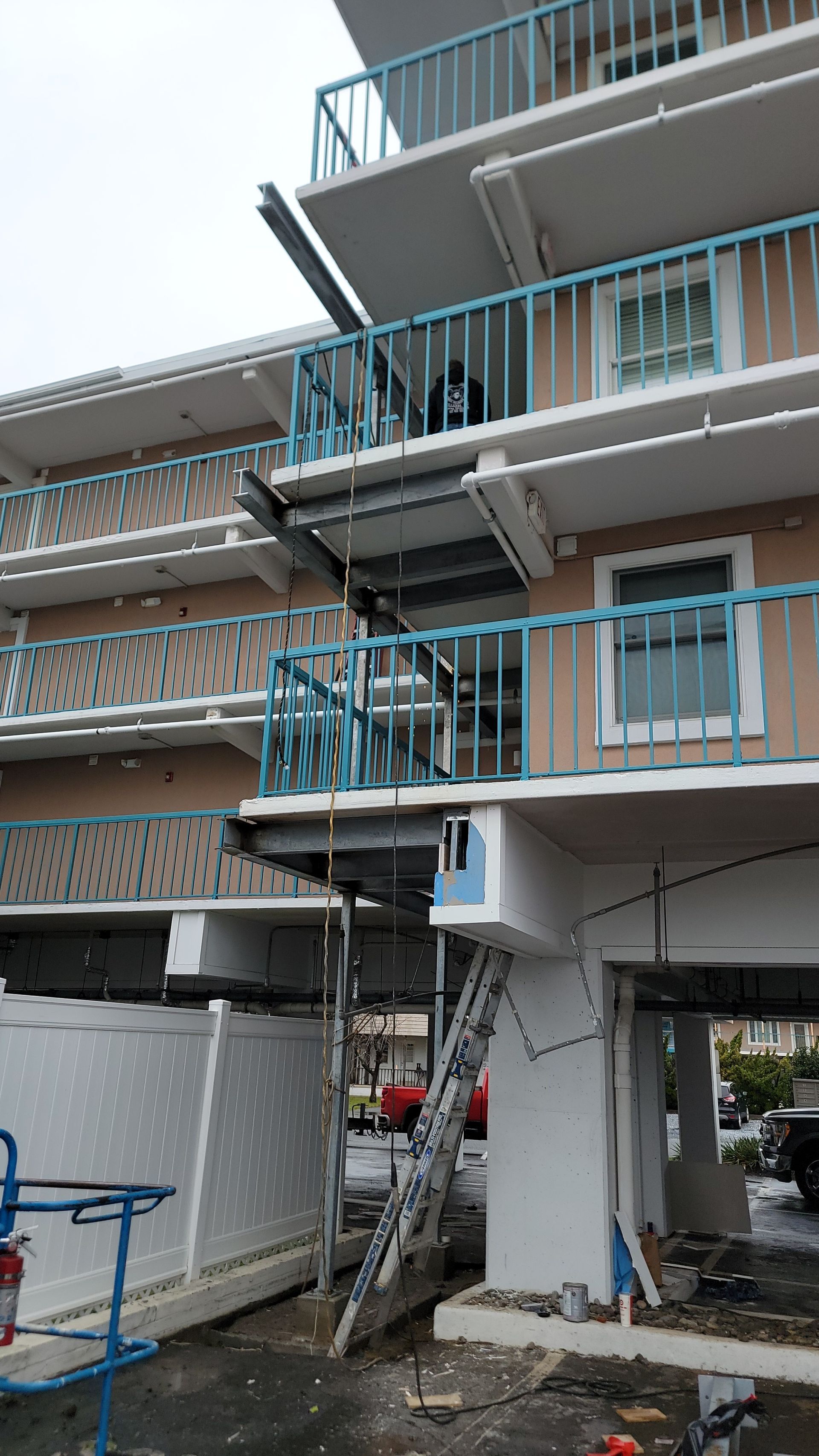Damaged building exterior with multiple levels, blue railings, and scaffolding. A ladder is propped against the building.