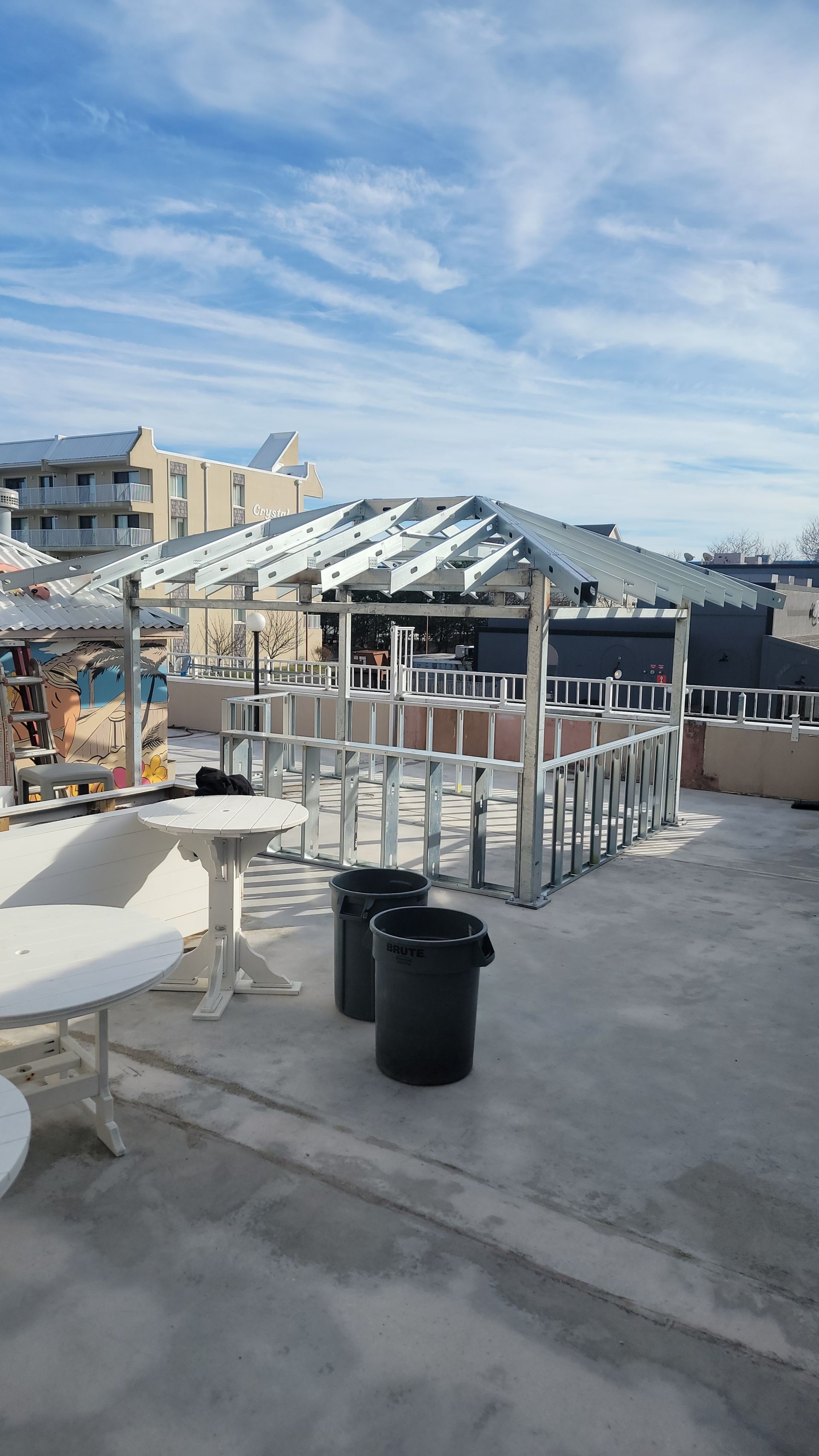Rooftop construction site with metal framing. Two trash cans and white tables. Cloudy sky in the background.
